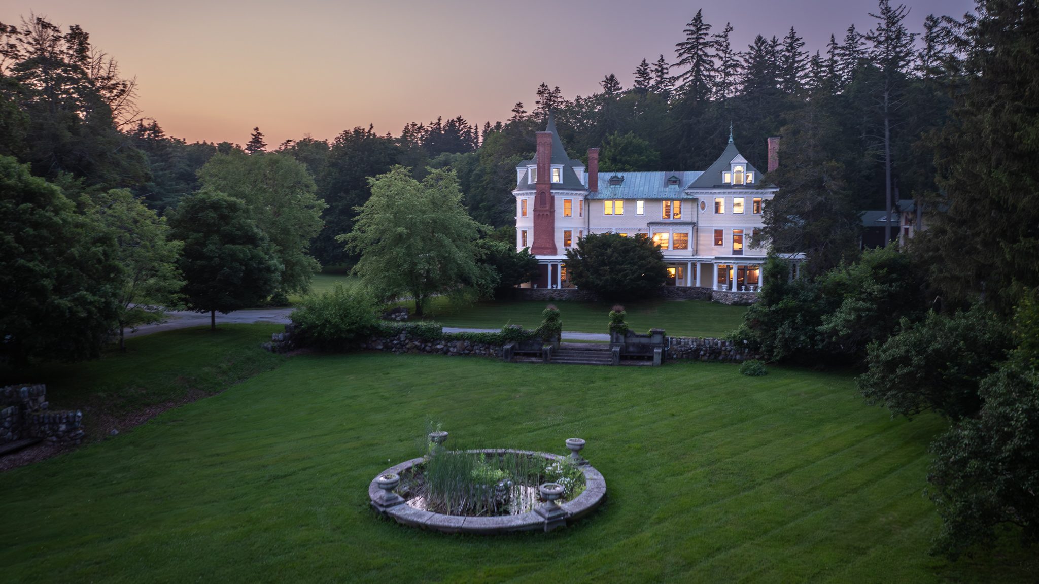 The round stone fountain in the foreground of the darkening lawn, the house glowing at the far end of it.