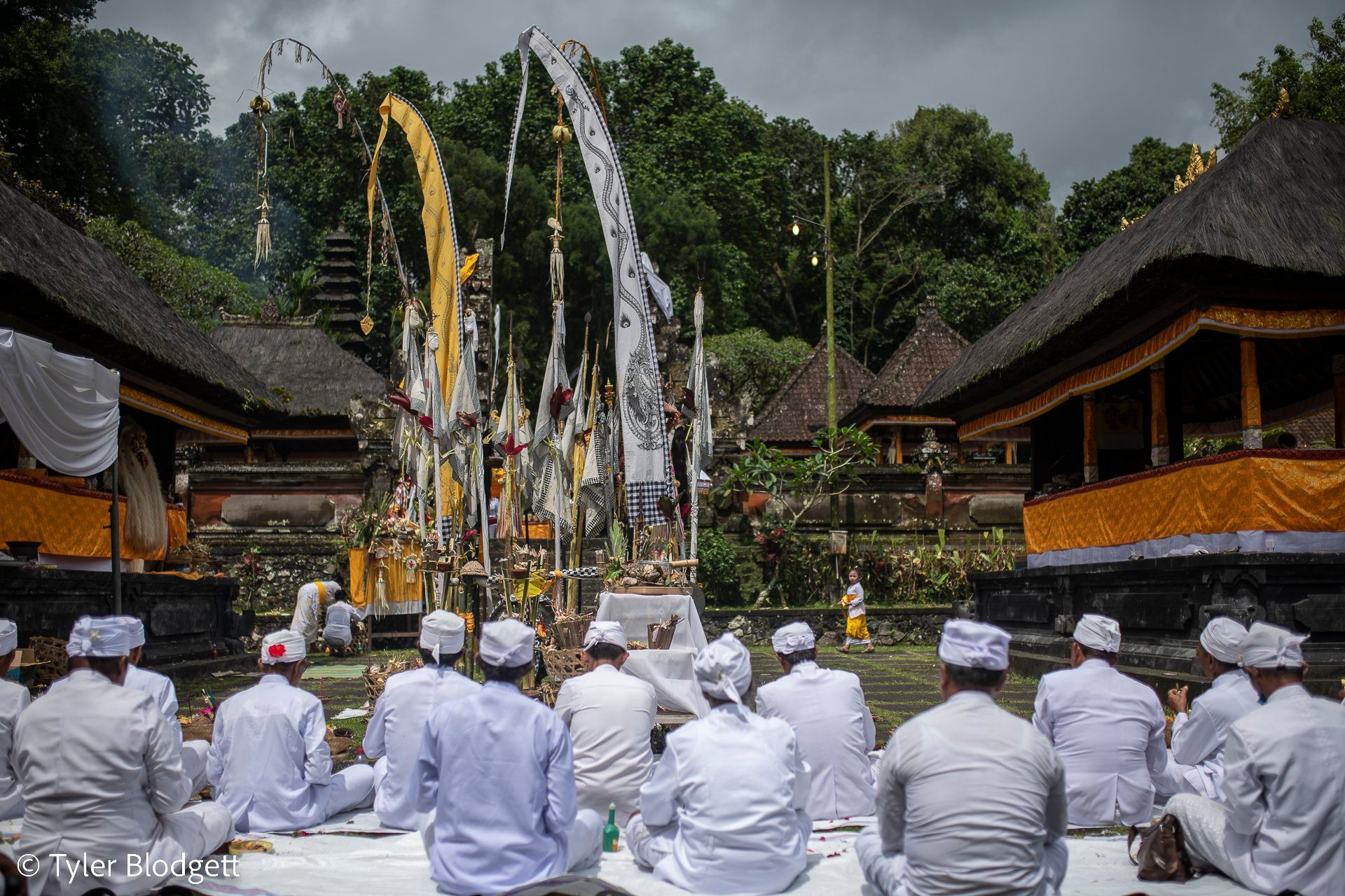 Rows of men sit in white facing the shrine, tall offerings of woven palm rising at its center.
