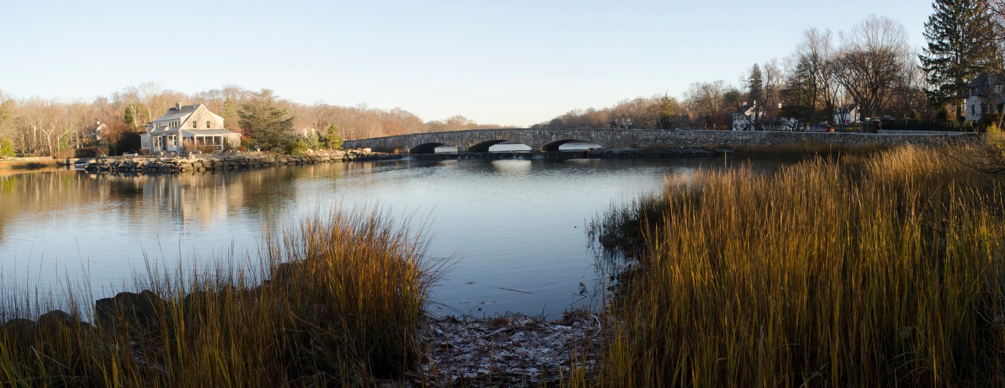 Marsh grass frames a stone bridge and a quiet house across the cove.
