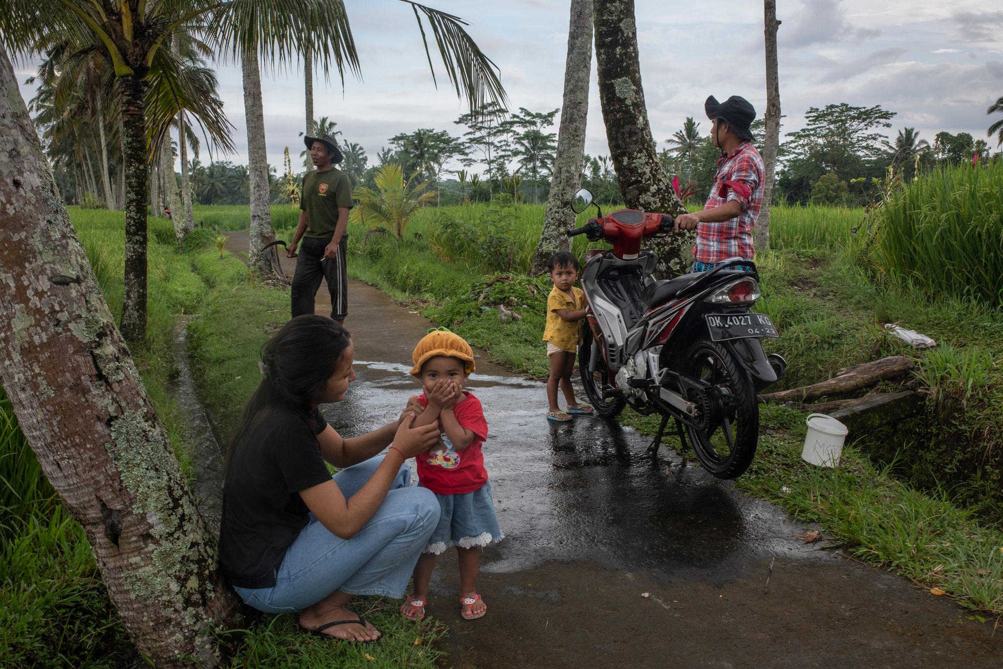 A mother crouches to talk with her daughter on a wet lane between the fields.