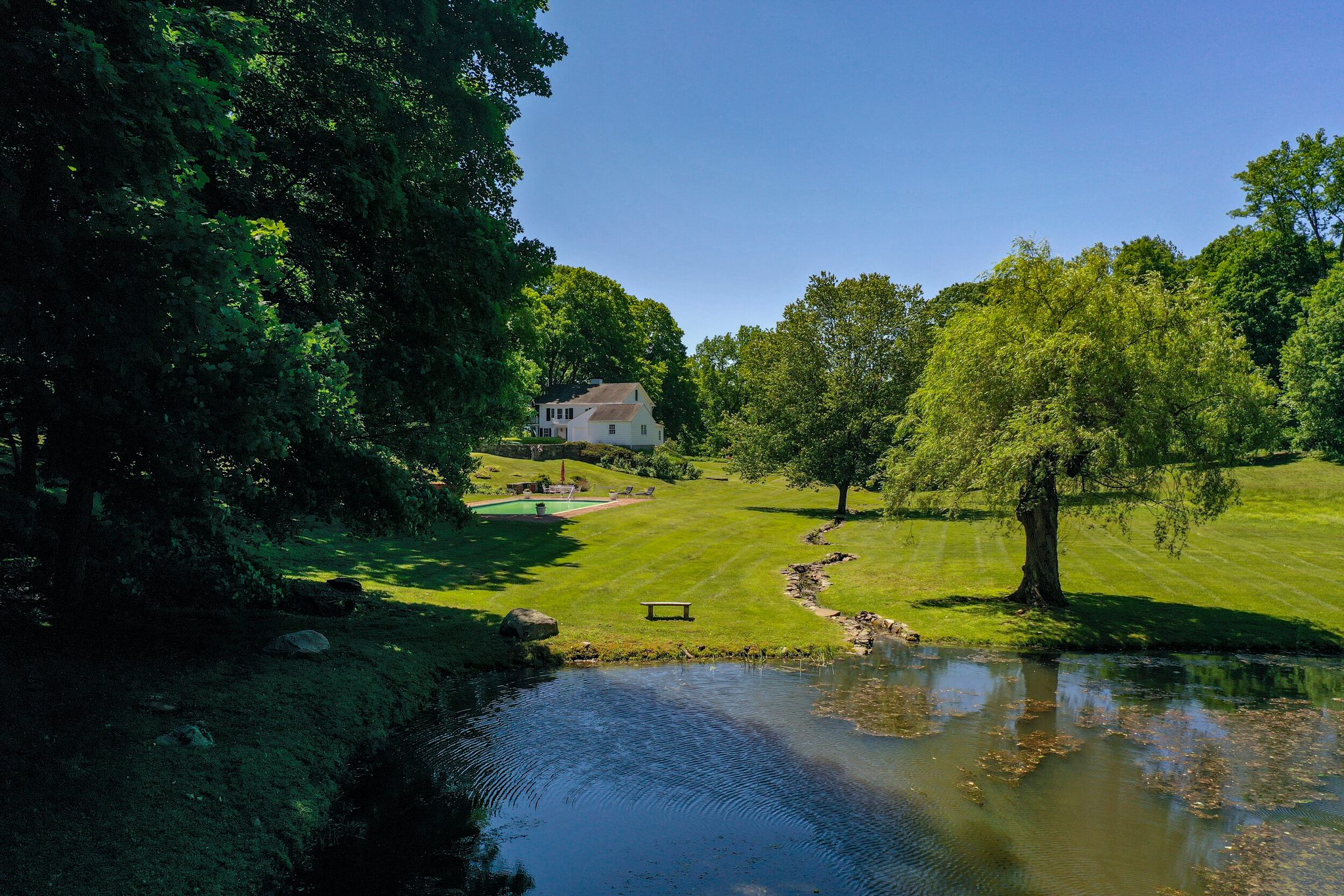 A stream winds through the lawn to the pond, a white house resting in the trees beyond.