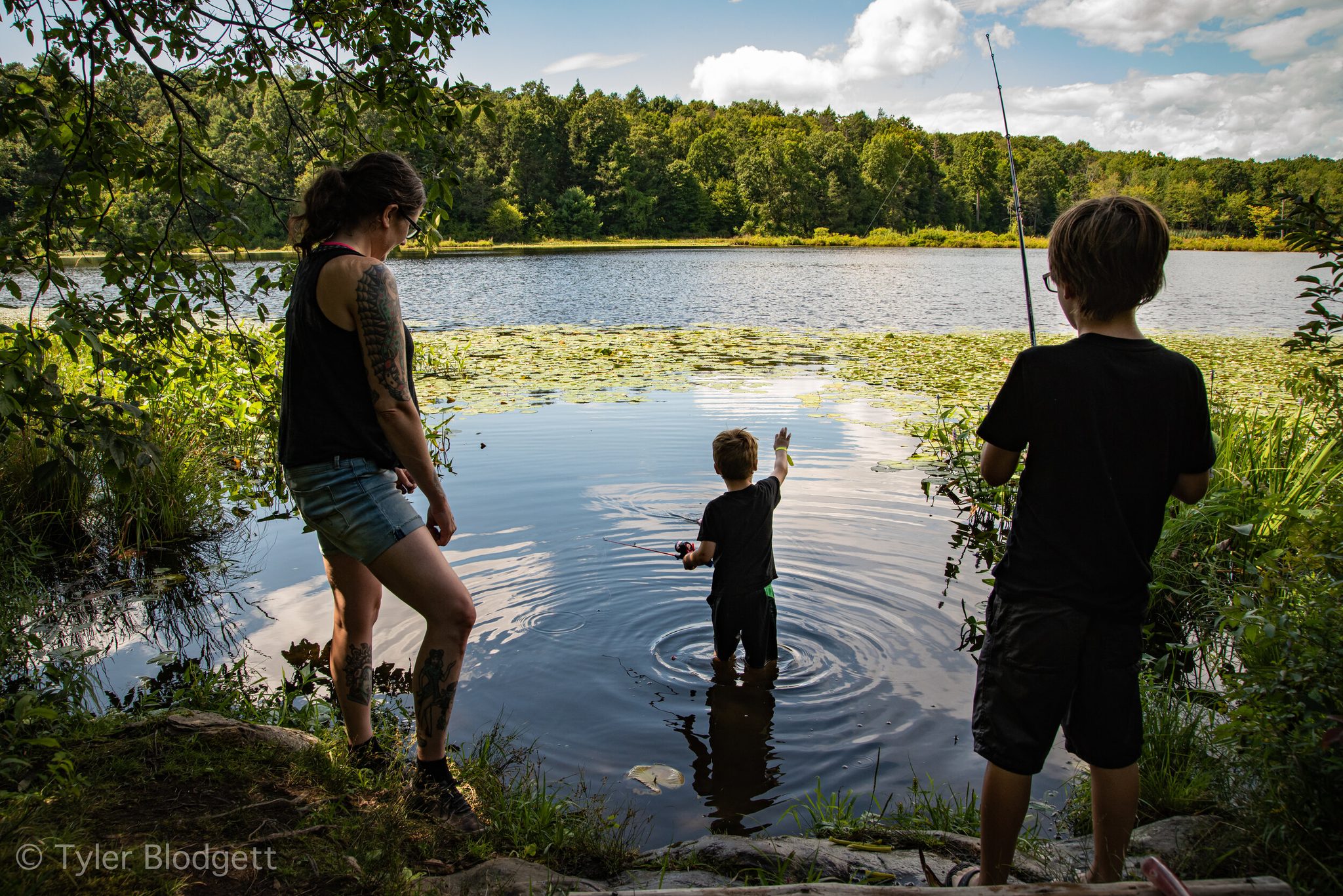 One boy wades in to his knees. Another casts from the bank, and the ripples spread through the lily pads.