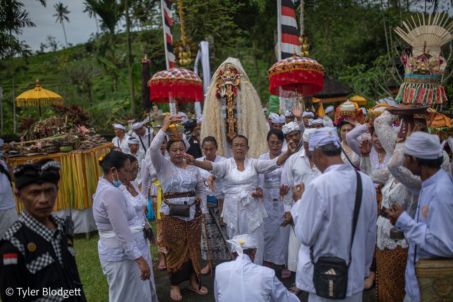 A woman in white dances at the center of the crowd, arms flung wide, while the masked figure looks on.