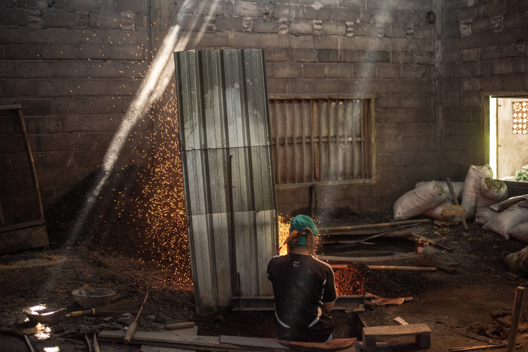 A shaft of light cuts through the smoke and sparks as a worker minds the forge.