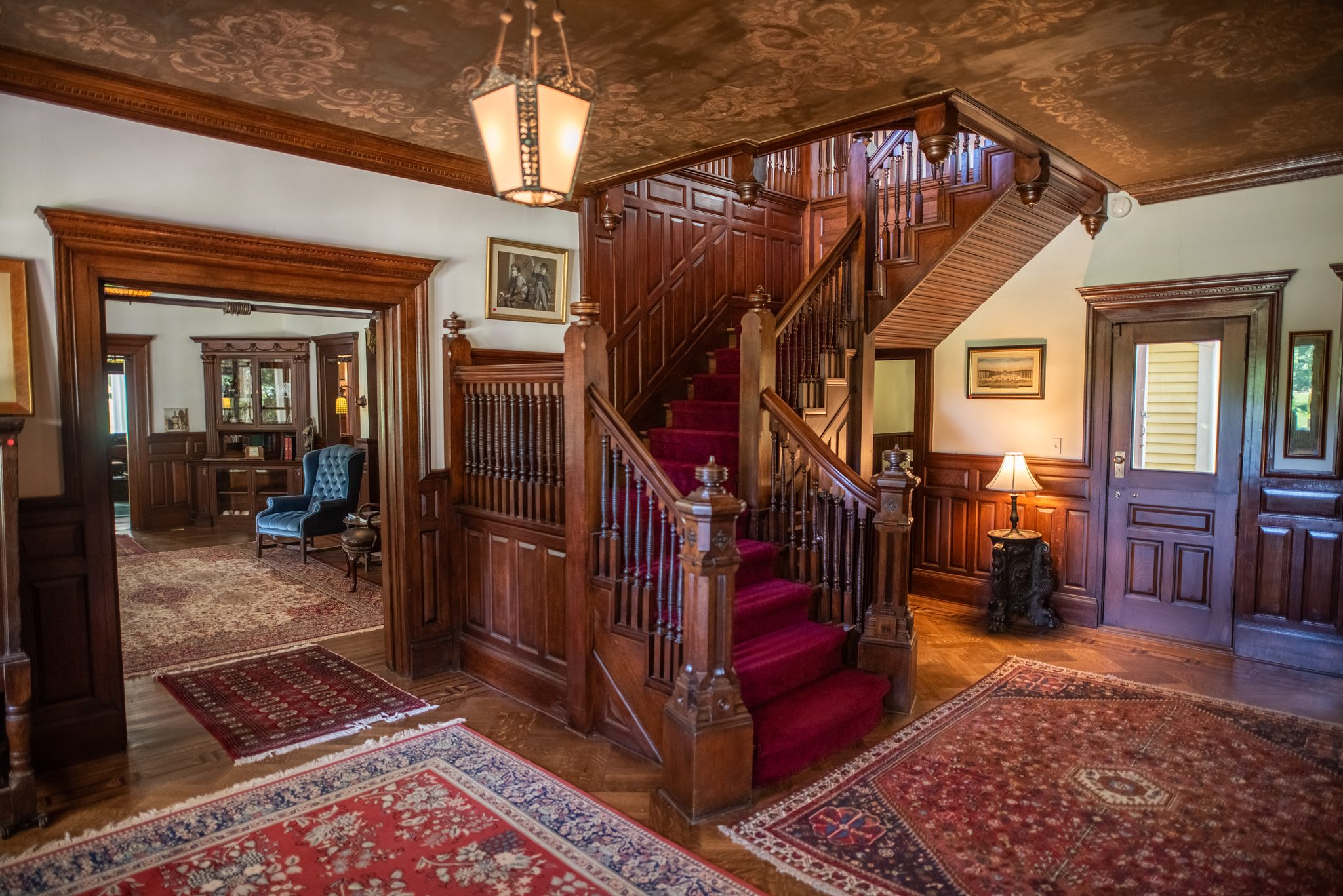 The front hall is all dark oak paneling, turned balusters and a red runner climbing the stair past a hanging lantern.