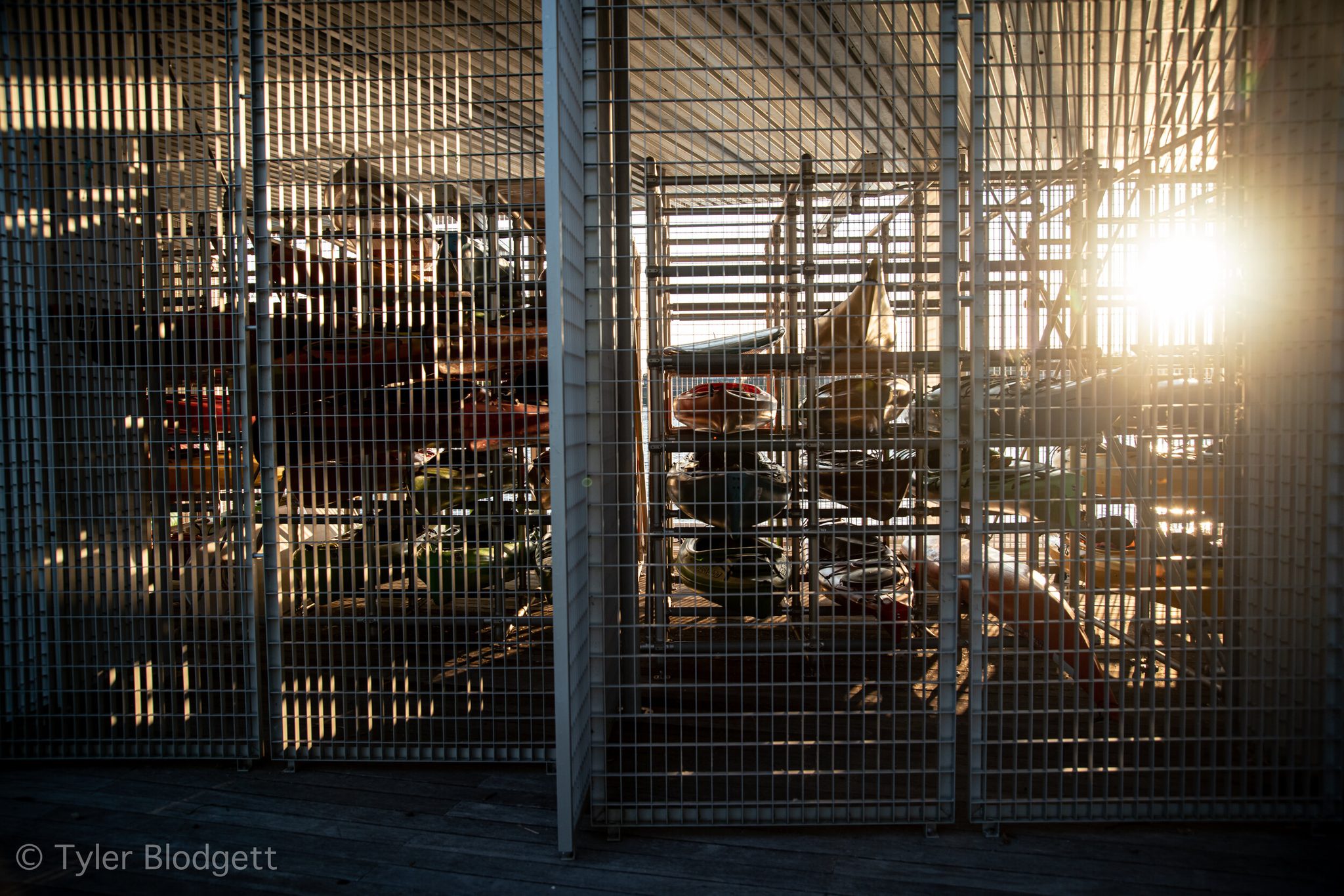 Kayaks rest in their steel racks behind the mesh, the late sun slipping through to find them.