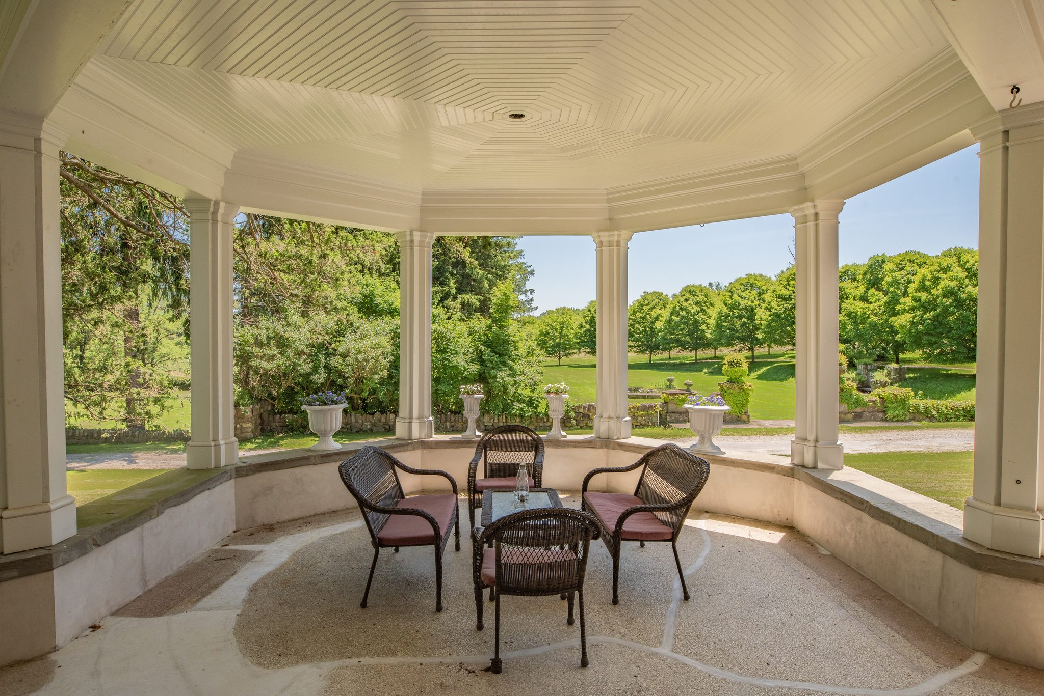 A round corner of the porch, wicker chairs under a beadboard ceiling and the lawn and orchard framed between white columns.