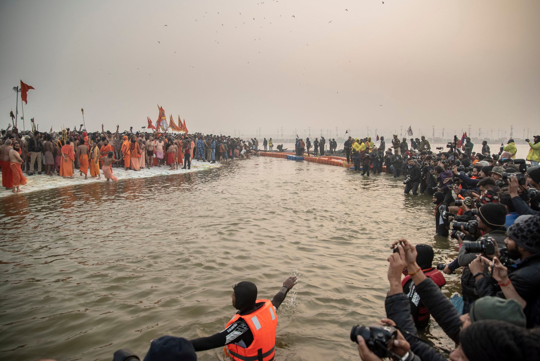Saffron flags crowd the far bank. In the water, a lifeguard waves toward shore as the cameras wait.