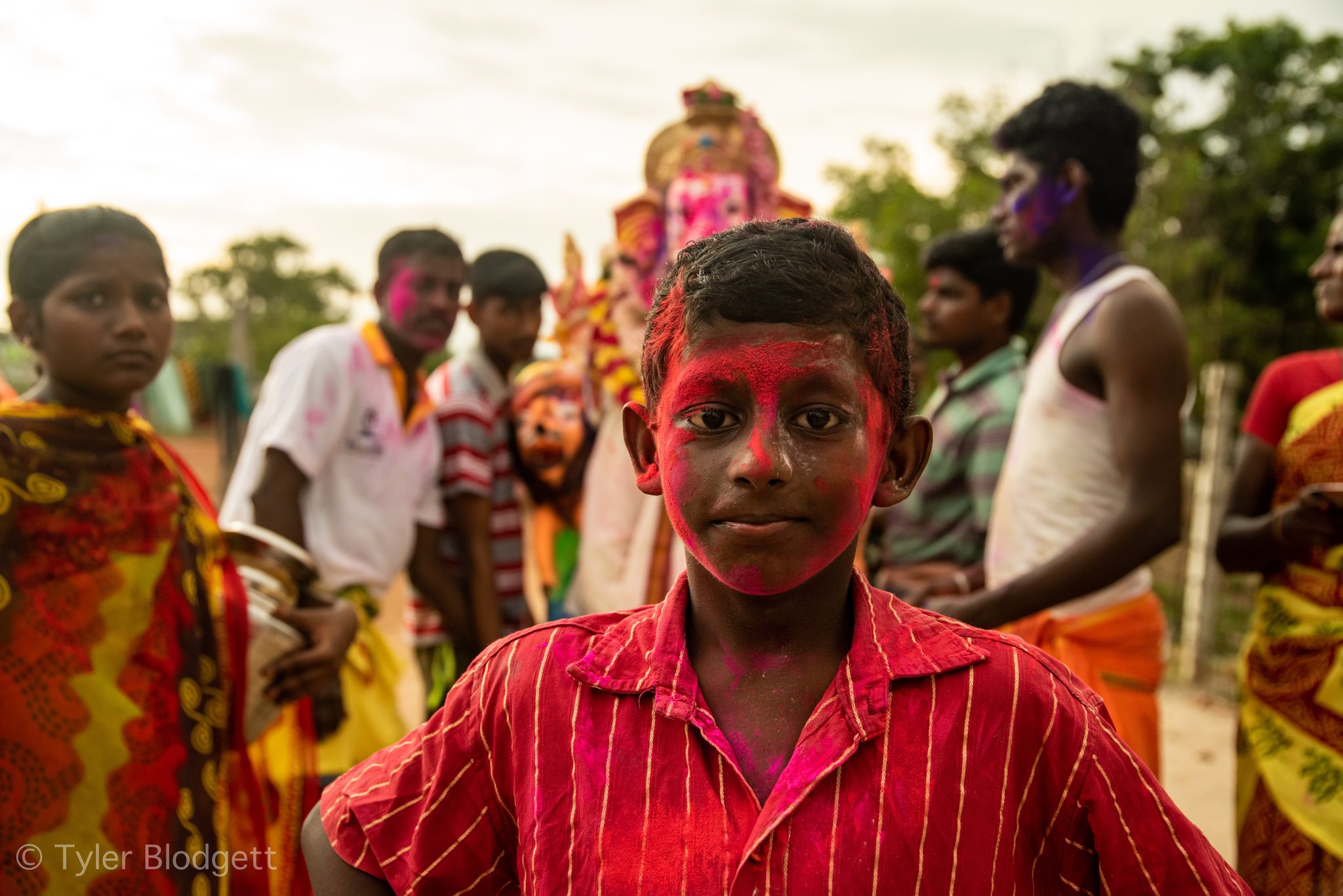 A boy with a face full of pink powder stands before the procession.