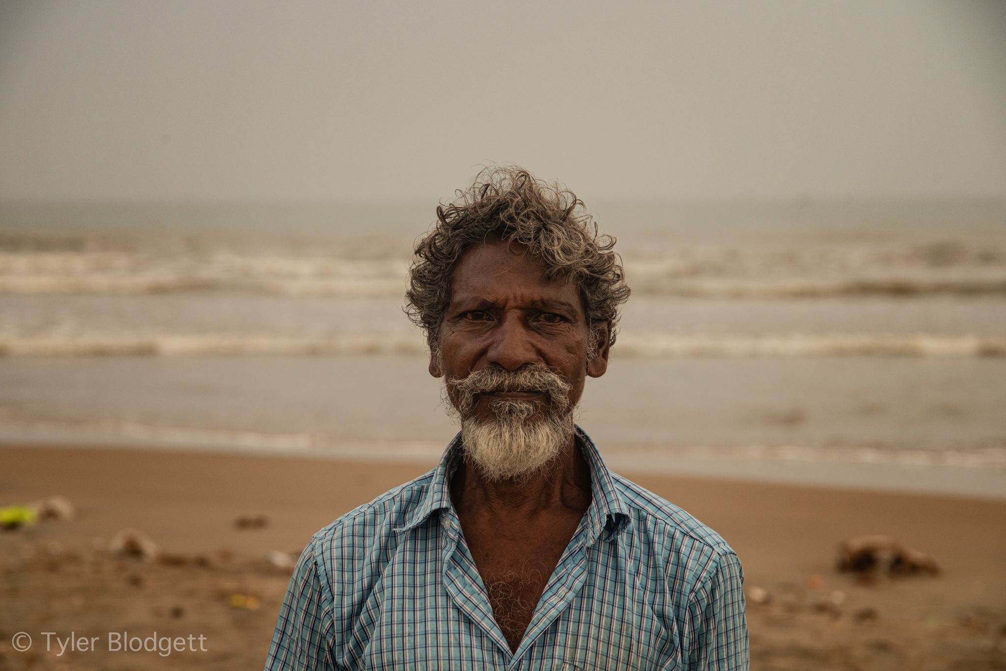 A fisherman with a white beard stands on the beach, the waves behind him.