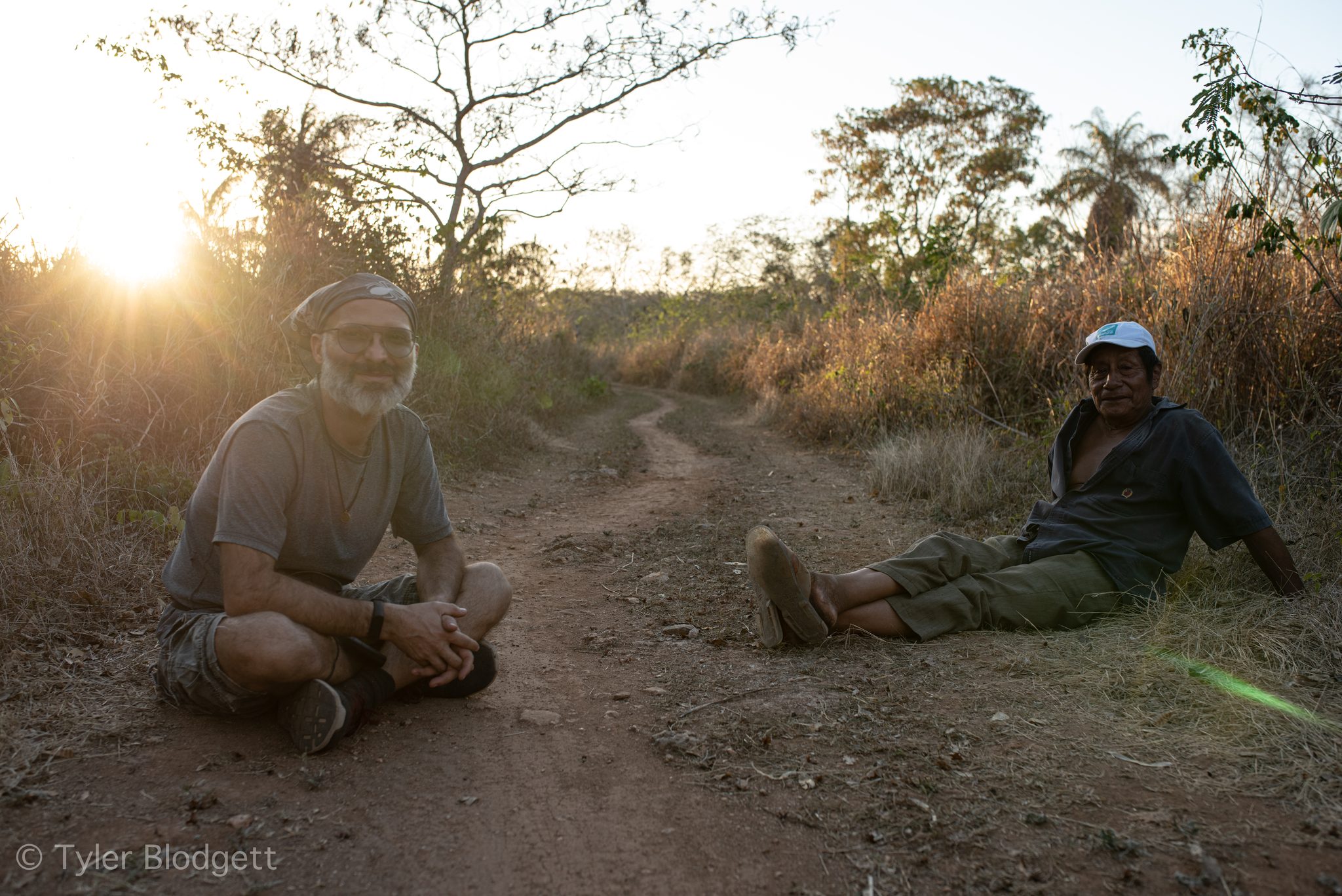 Two men sit in the dirt road as the sun sets behind them.