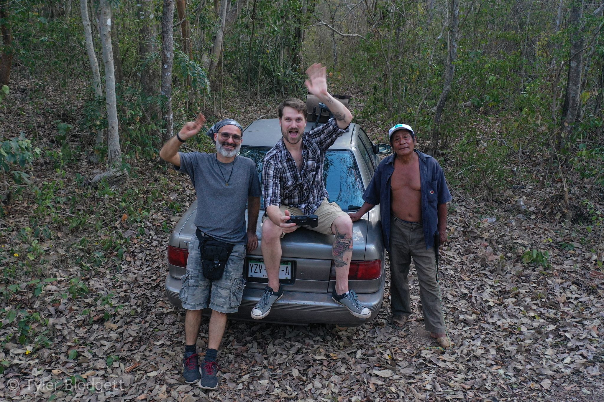 Three men wave from the trunk of a car in the forest.
