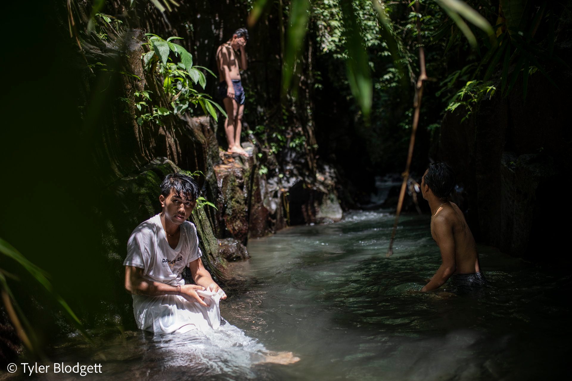 One boy climbs the rock to jump. His friend sits waist deep in the green pool, waiting to see if he will.