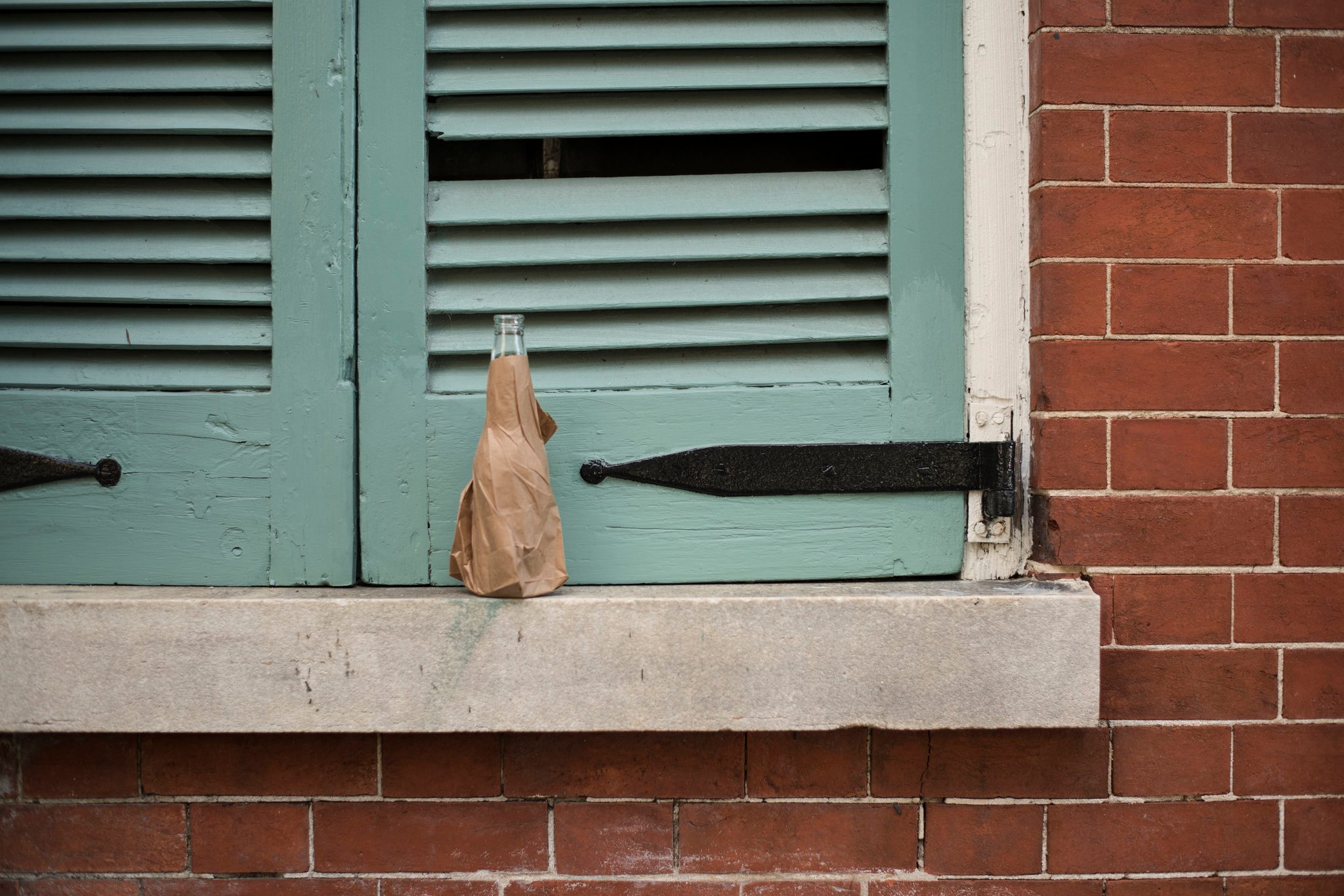 A bottle in a paper bag rests on the sill beneath green shutters.