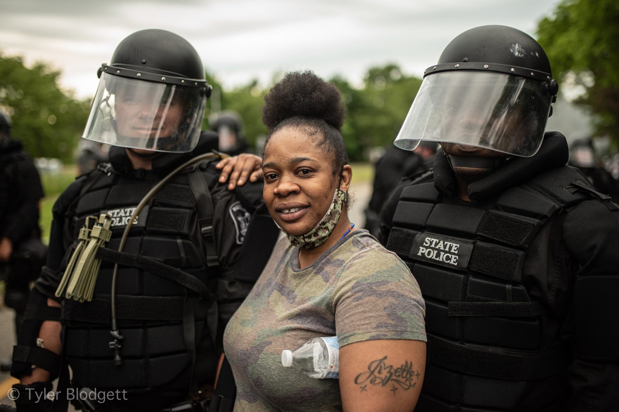 A woman smiles between two troopers in riot gear, one of them resting a hand on her shoulder.