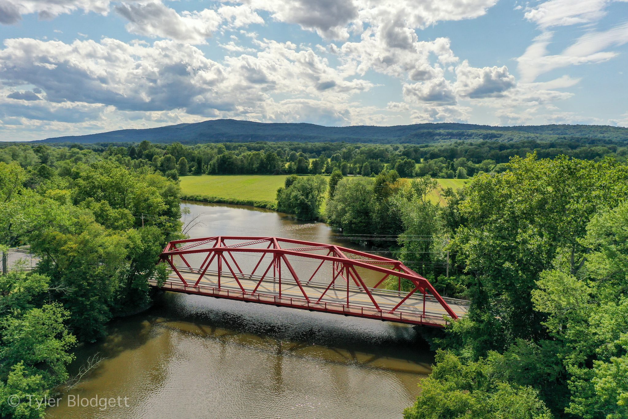 A red truss bridge spans the river between fields and forest, the hills rolling away to the horizon.