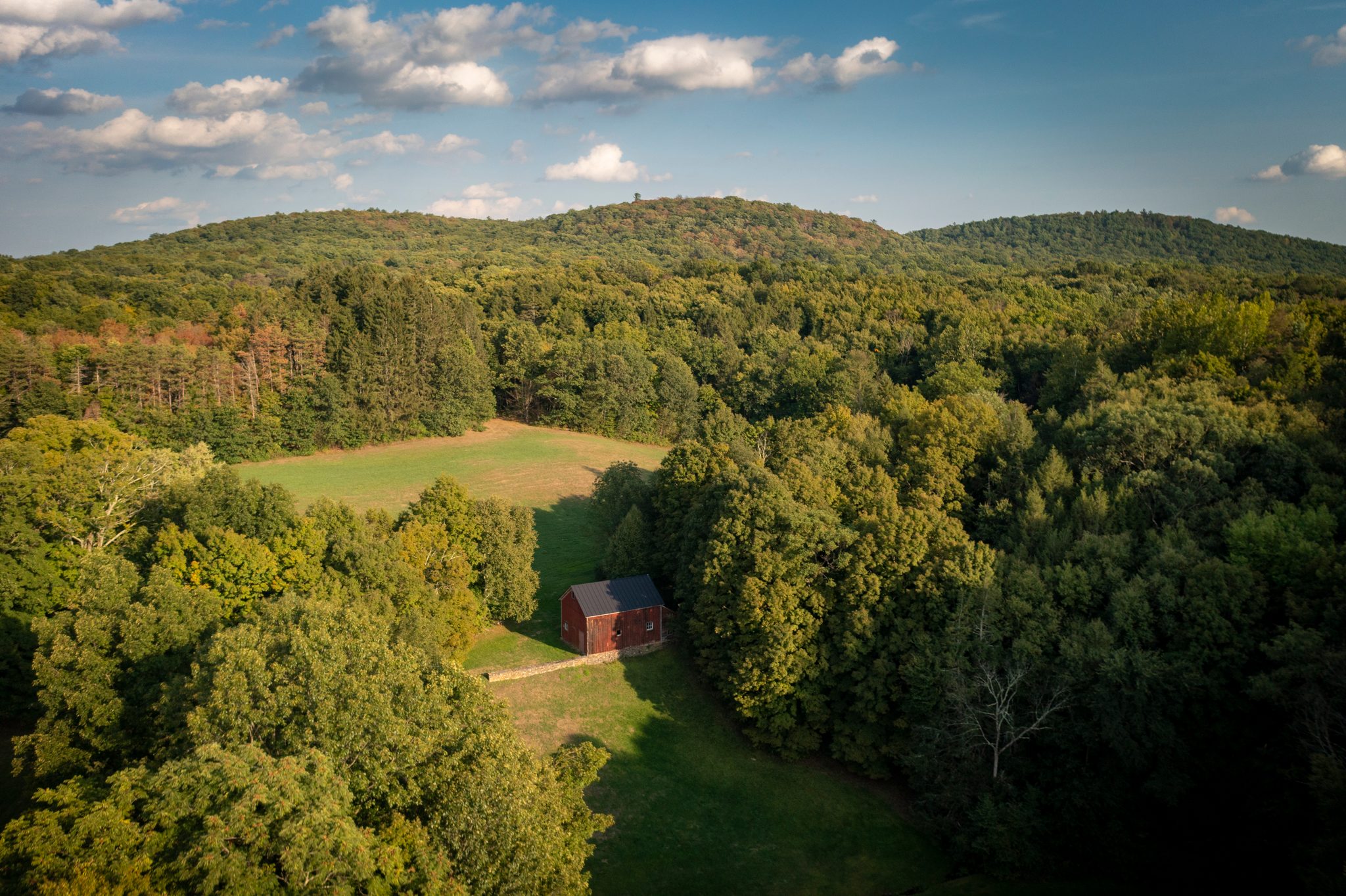 The red barn sits at the bottom of its meadow, forest on every side and the hills beyond.
