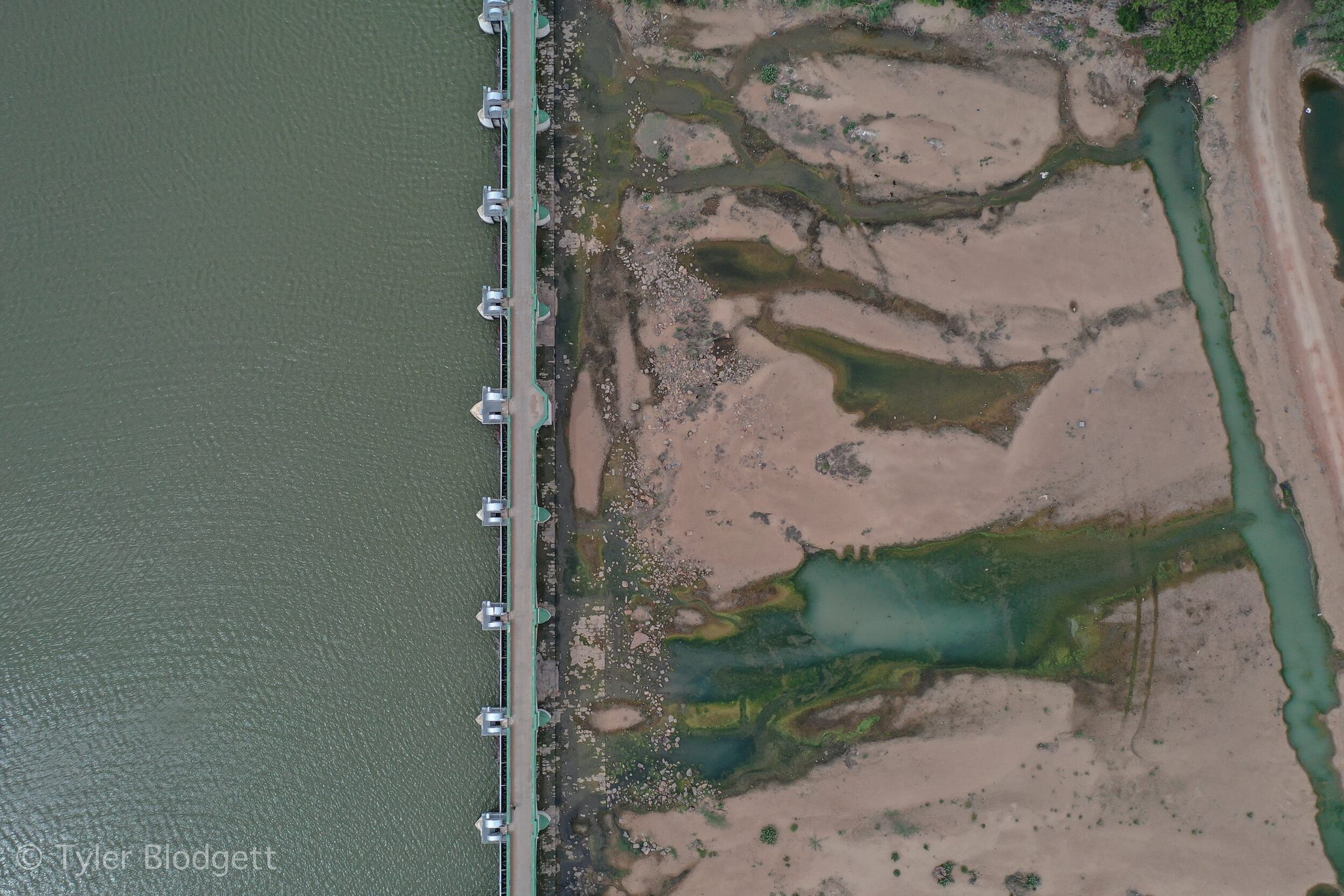 From above, a barrage holds the river on one side and leaves the other to sand and green pools.