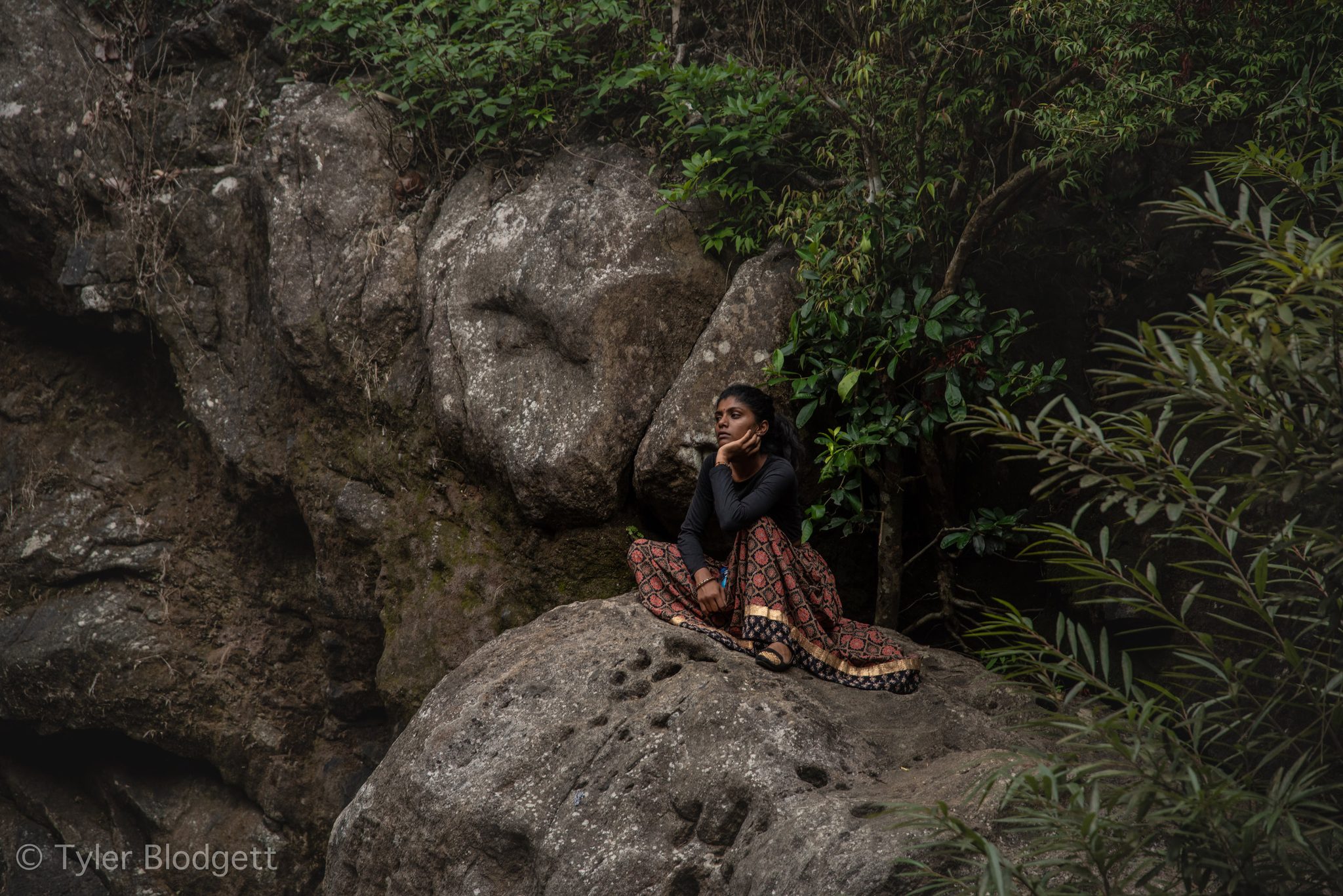 A young woman rests her chin in her hand on a mossy boulder, the forest quiet around her.