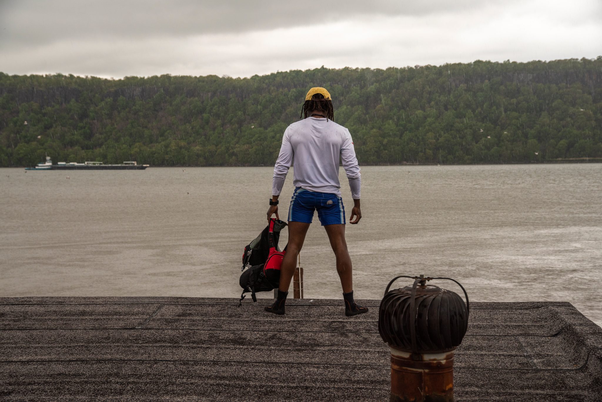 Chev stands on the end of the dock with his gear at his feet, looking out over flat grey water.