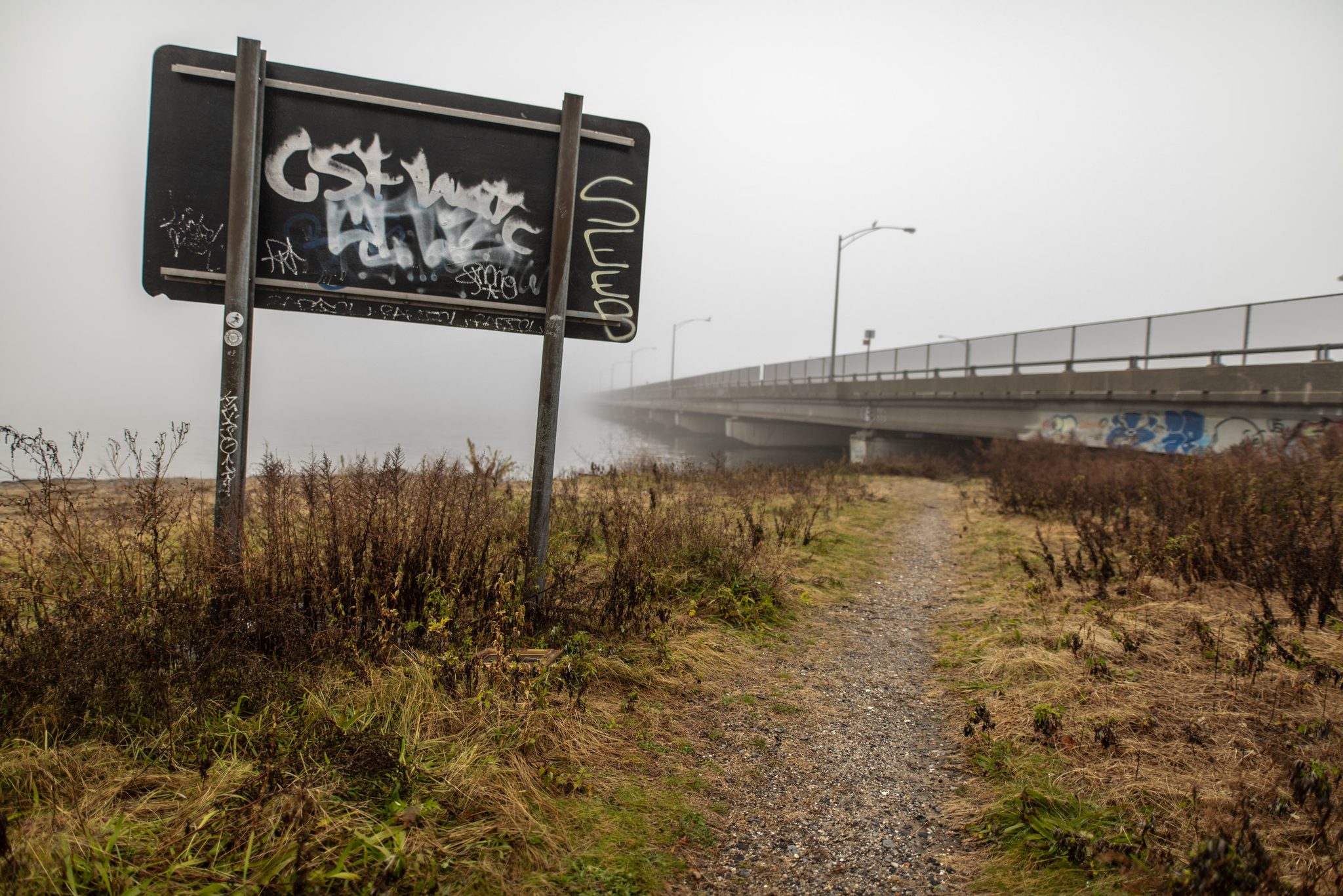 A path runs off into the fog beside a graffitied sign and the ghost of a bridge.