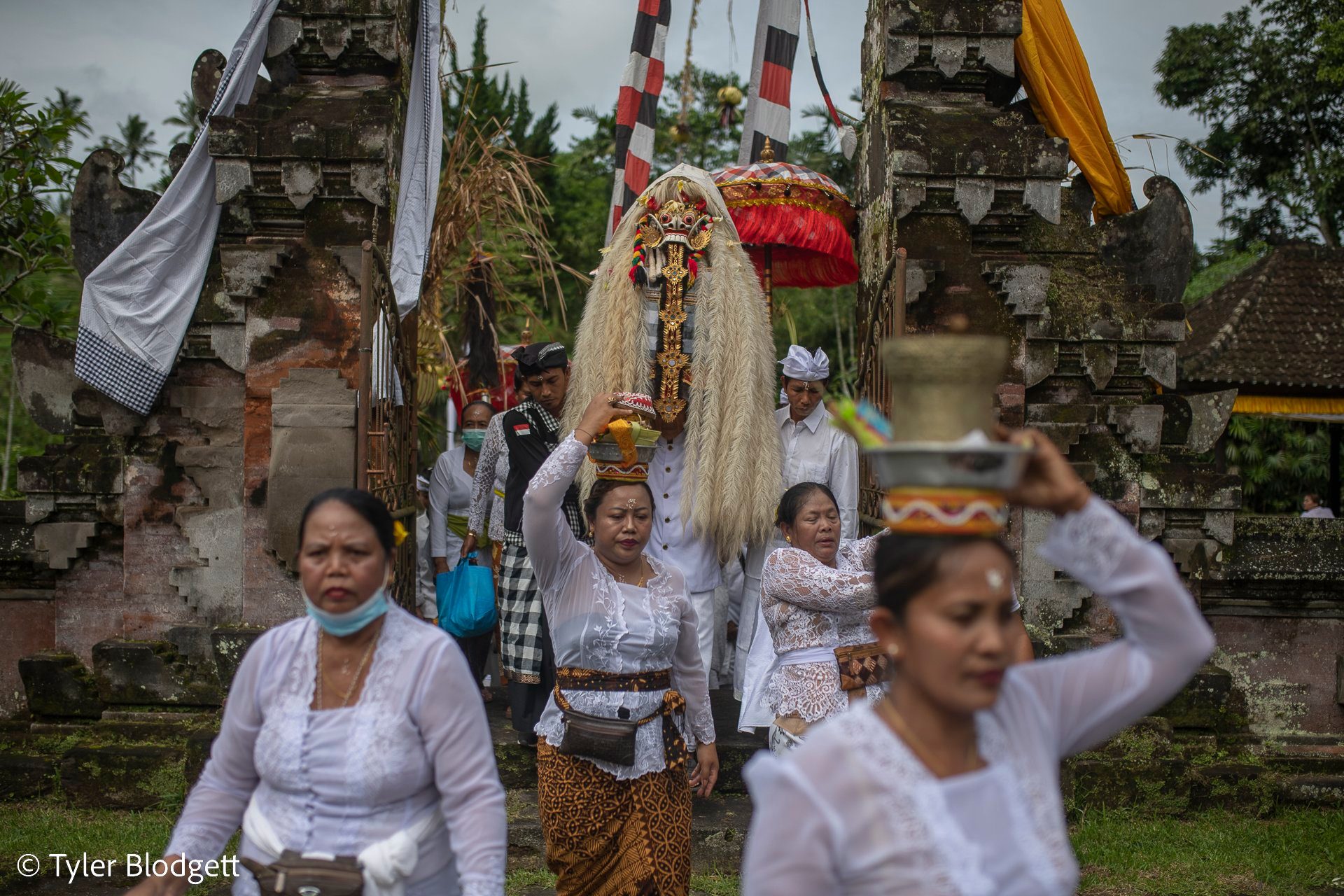 Women carry offerings through the split stone gate, the masked figure close on their heels.