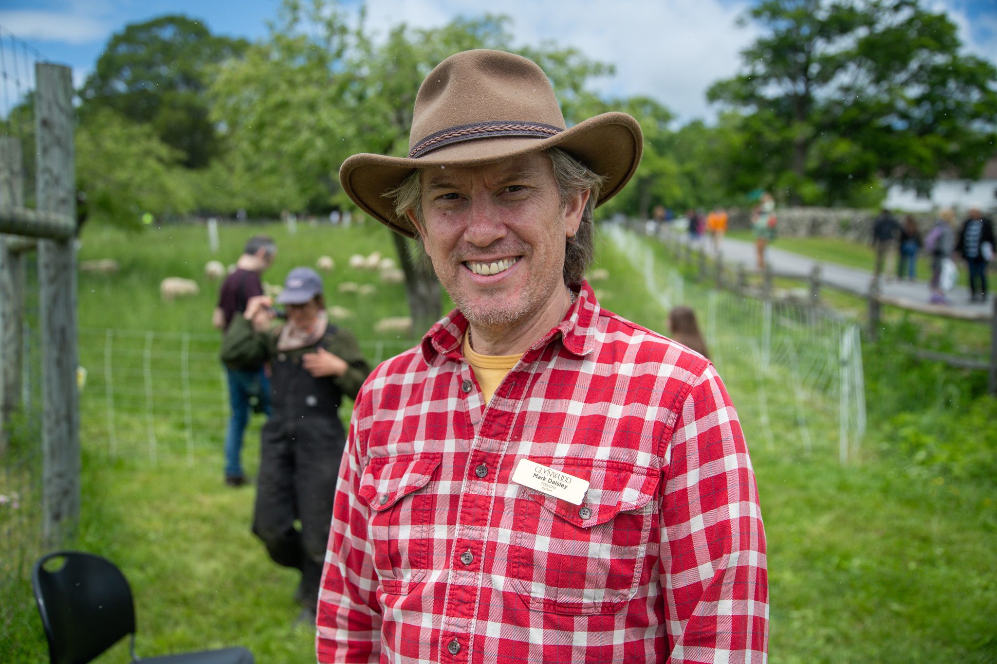 A farmer in a wide hat and red plaid smiles at the edge of the sheep pasture.
