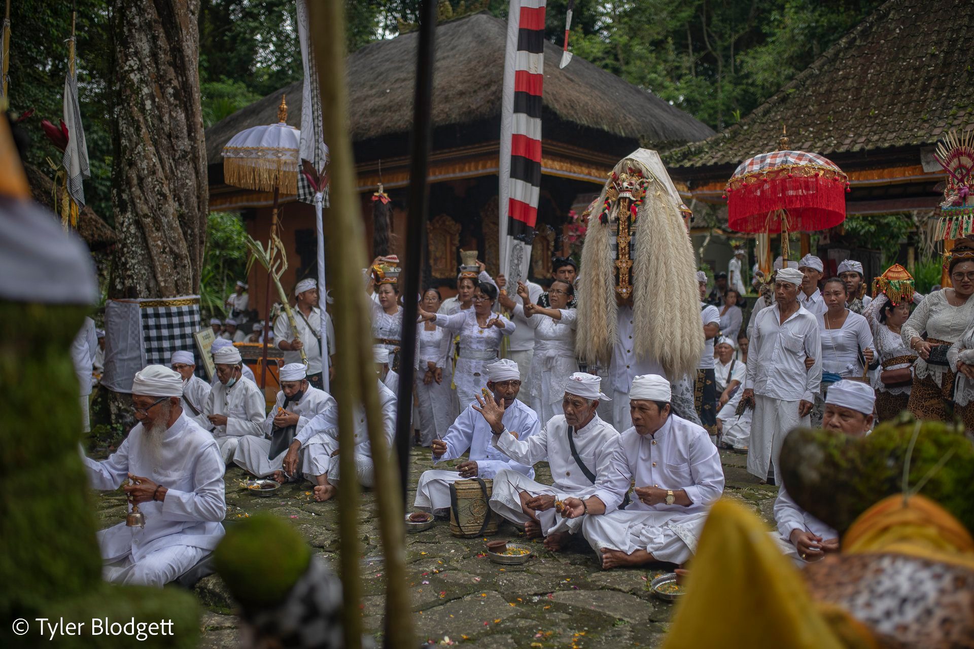 Men sit cross-legged in prayer, the masked figure standing among the parasols.