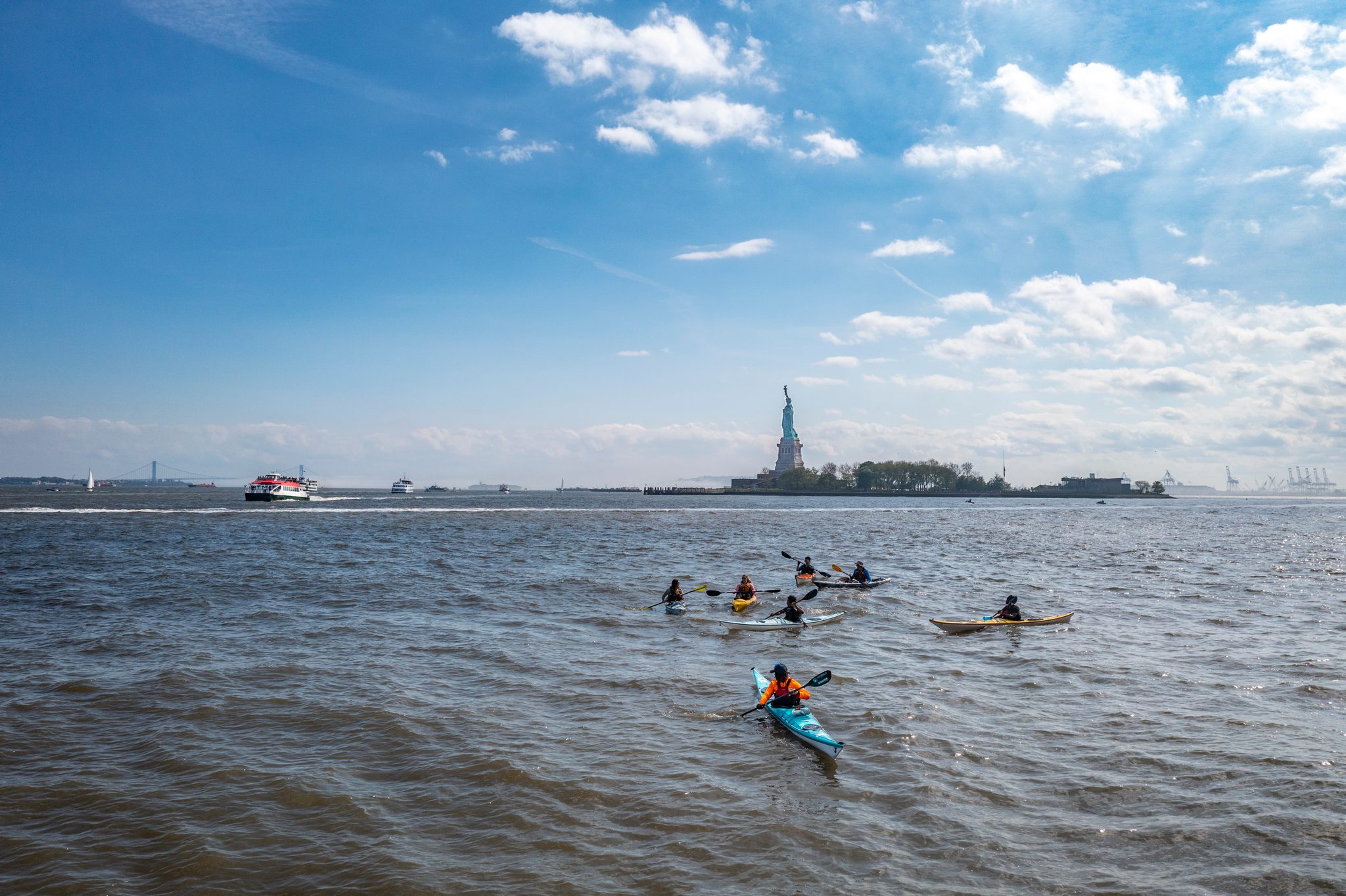From the drone, a scatter of kayaks crosses the harbor with the Statue of Liberty ahead under a big blue sky.