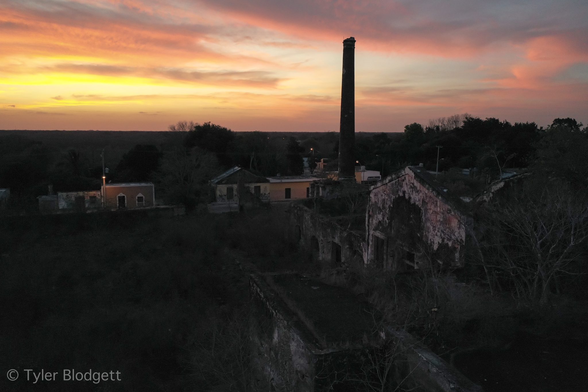 An old chimney stands against a blazing sunset over the rooftops.