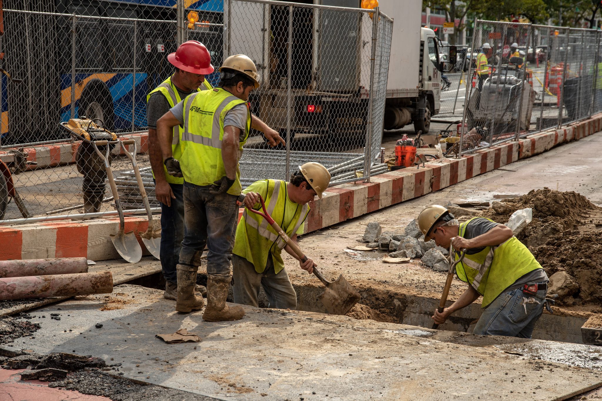 Hard hats bent to the trench, a crew digs out the sidewalk.