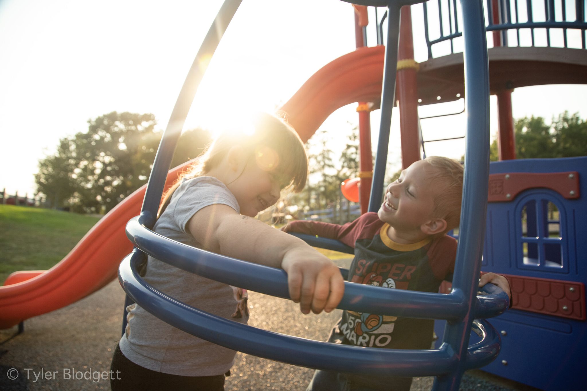 Two boys hang off the playground climber, the late sun pouring through the bars.