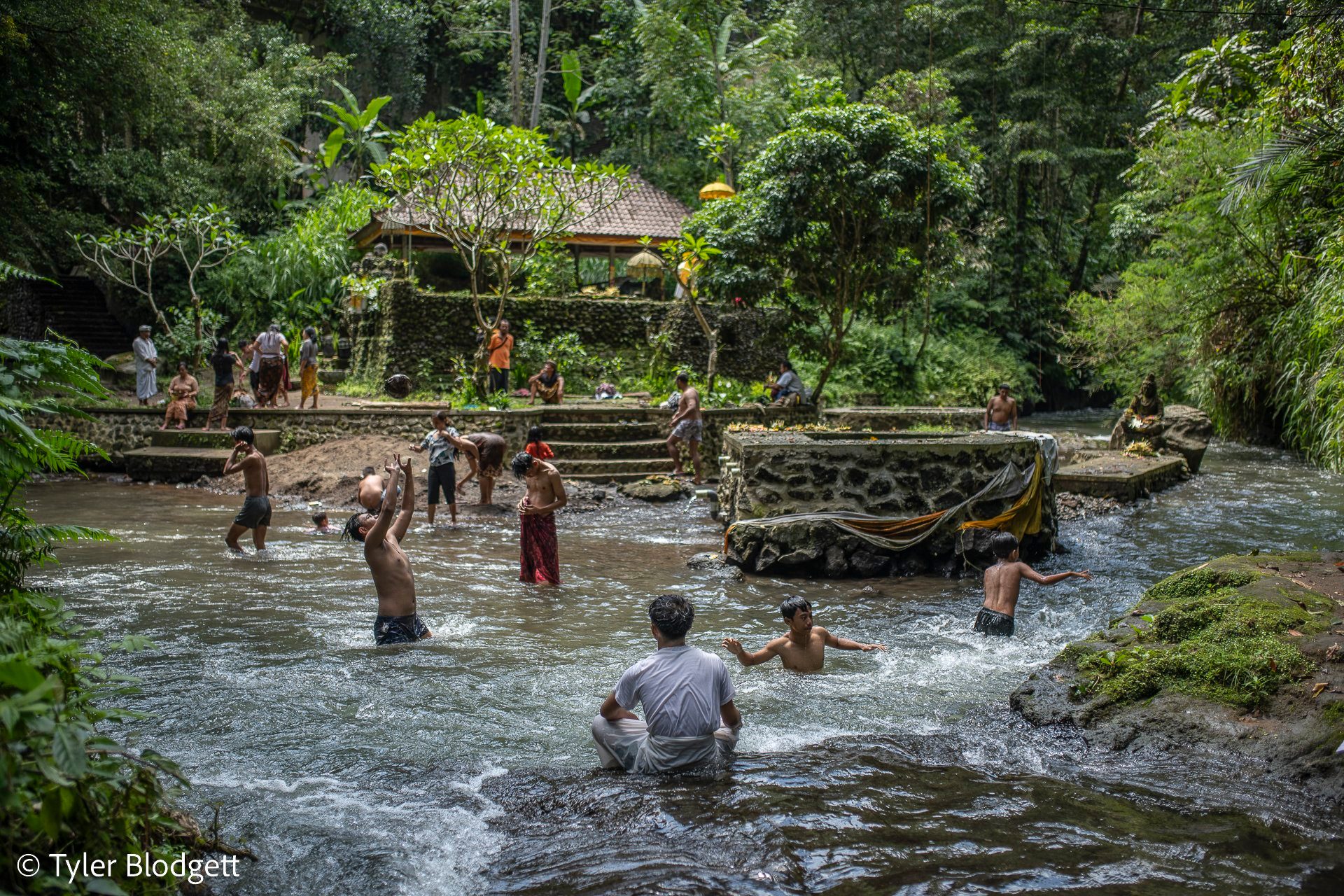 Families wade and bathe in the river below the temple steps, the jungle closing in on every side.
