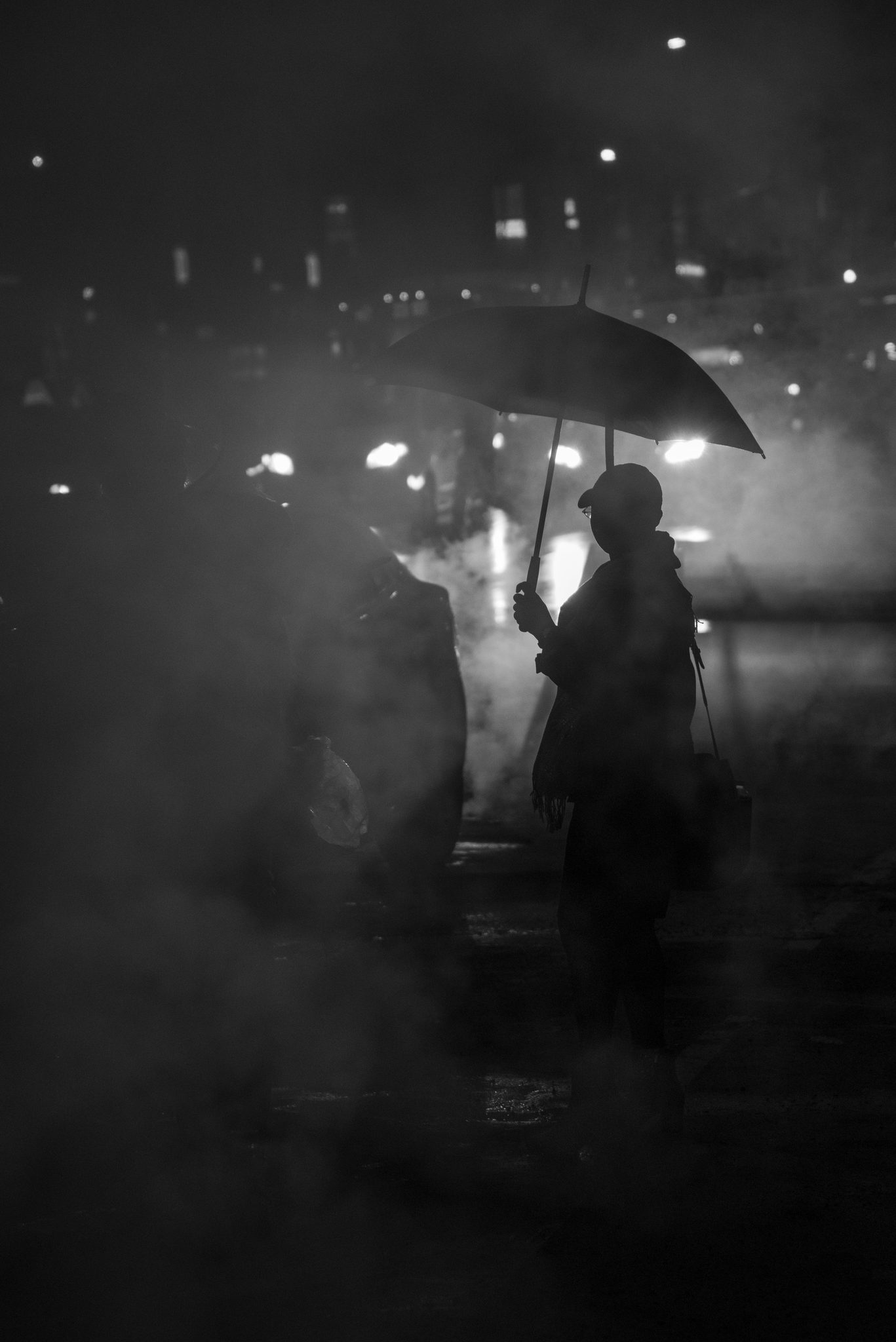 In black and white, a figure under an umbrella waits in the rising steam.