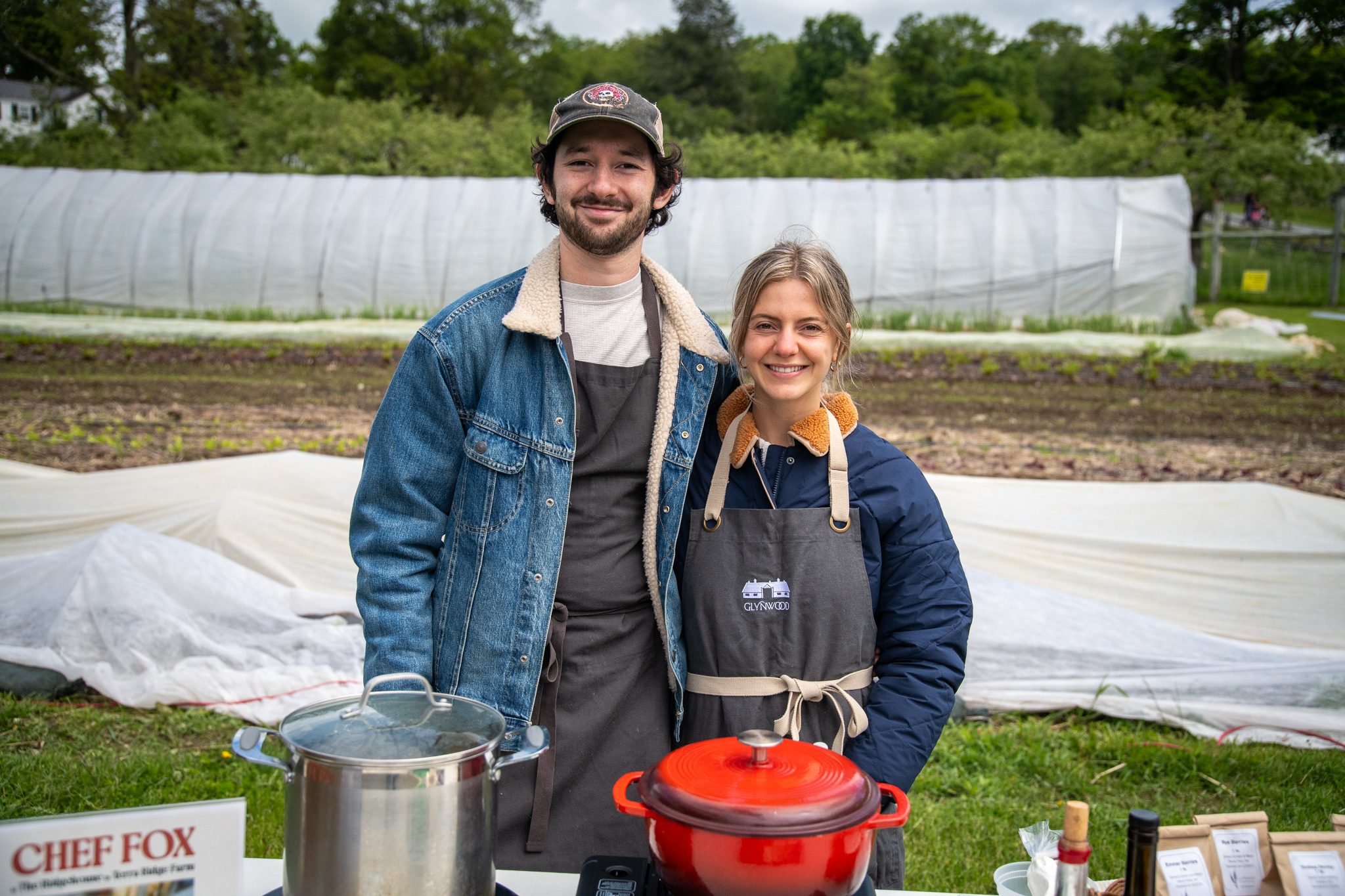 The two cooks behind the demo table pose with their pots in front of the hoop houses.