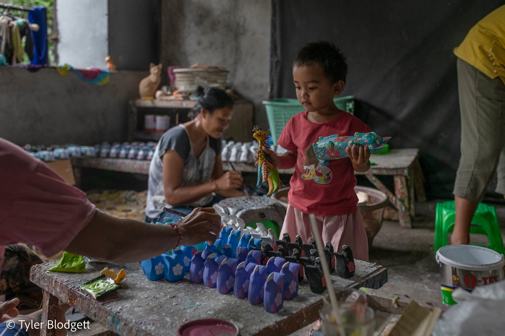 A child holds up a freshly painted carving. Around the table, the painting never stops.