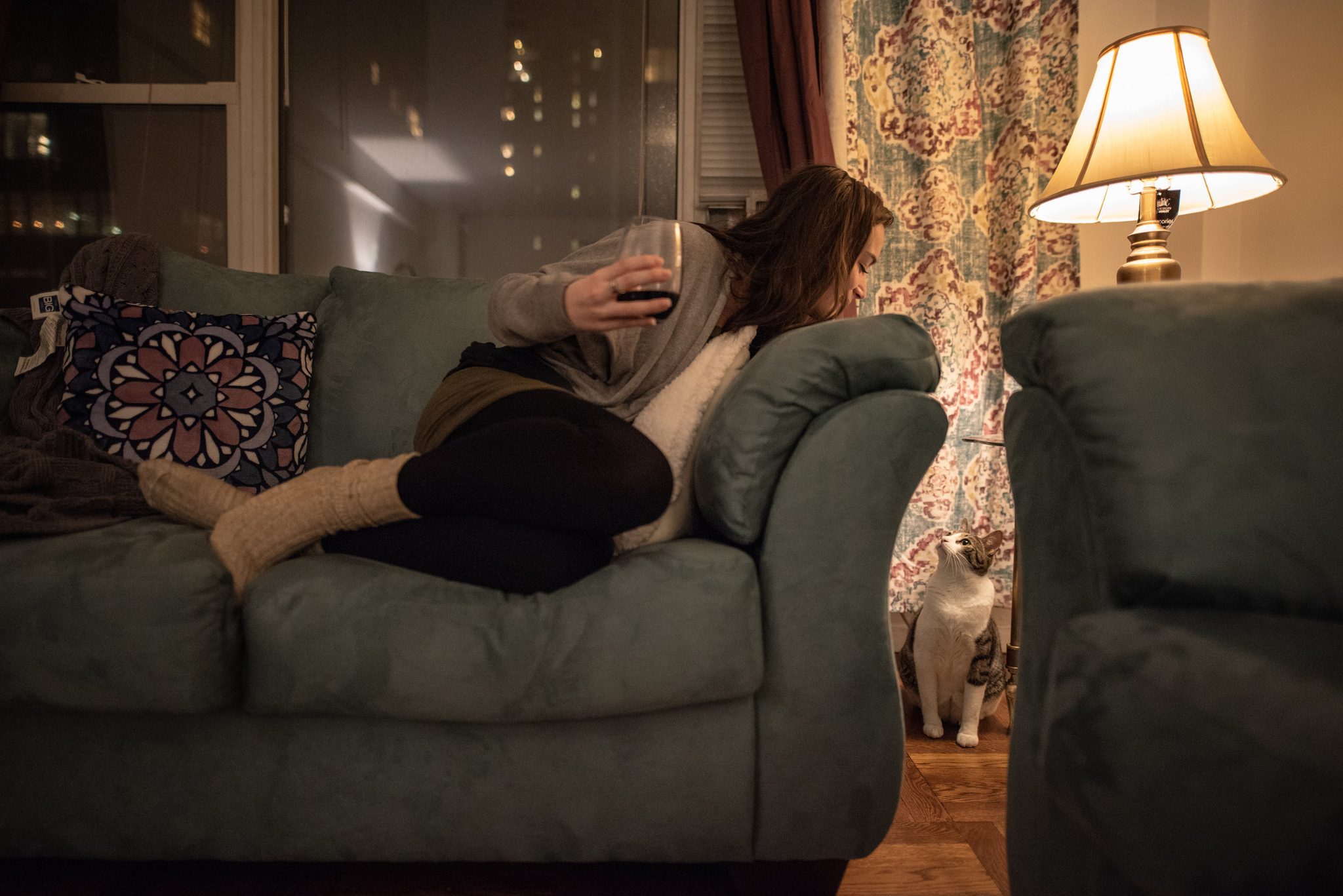 She curls into the couch with her phone. The cat keeps watch below the window, and the city glows behind the curtains.