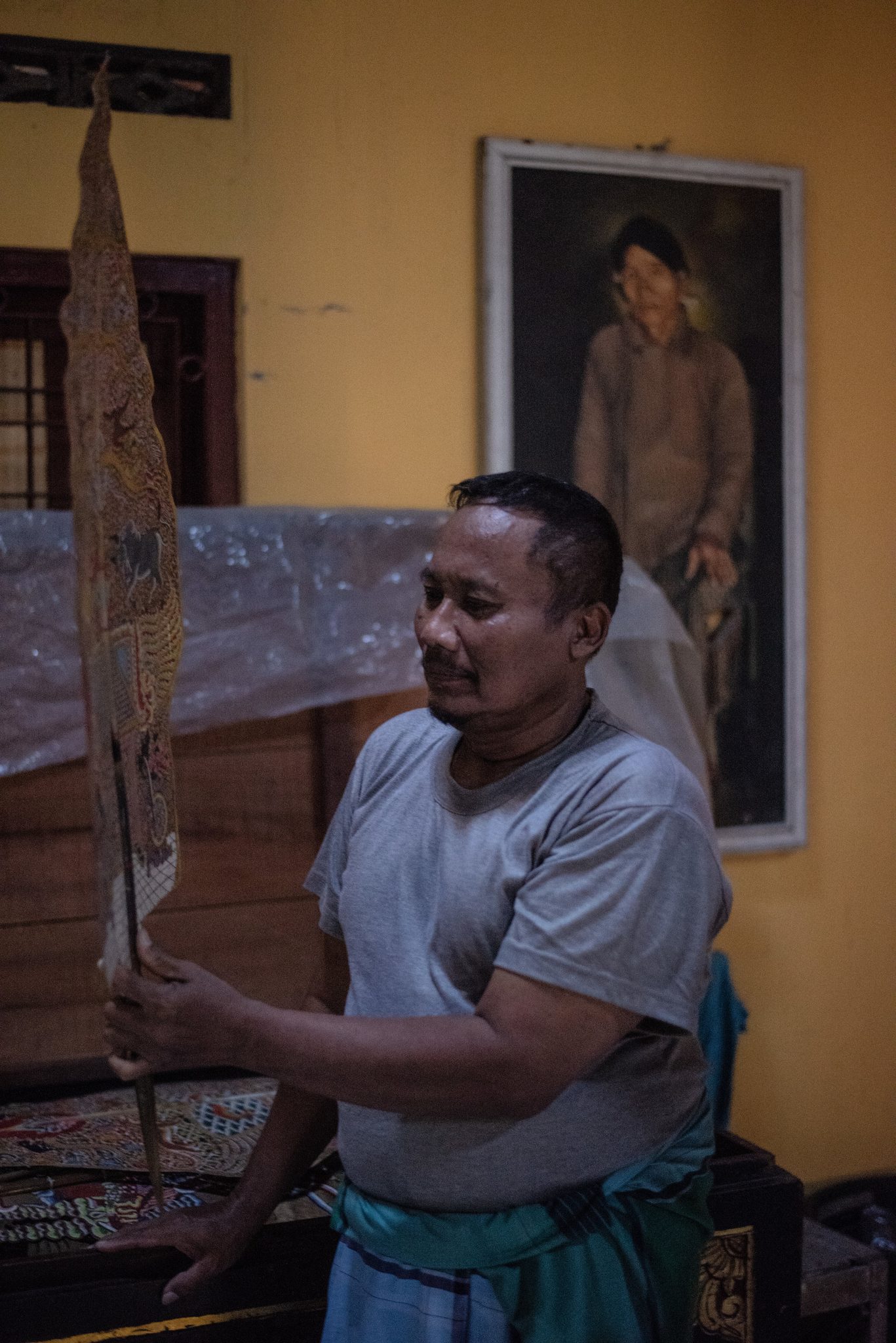 He holds the puppet upright and studies it, a portrait of an elder watching from the wall.
