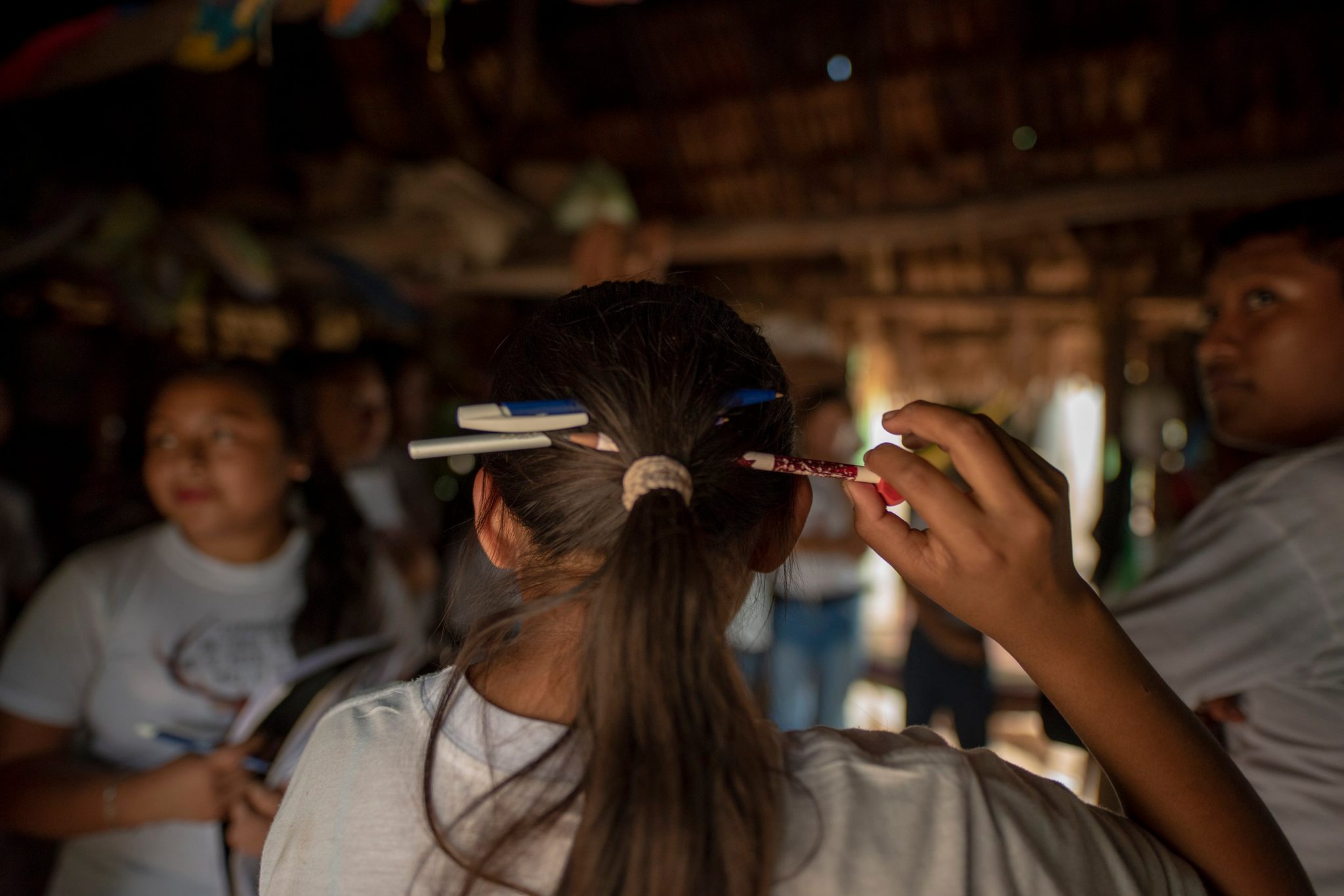 Pens tucked into her ponytail, a girl leans in to the lesson under the thatched roof.