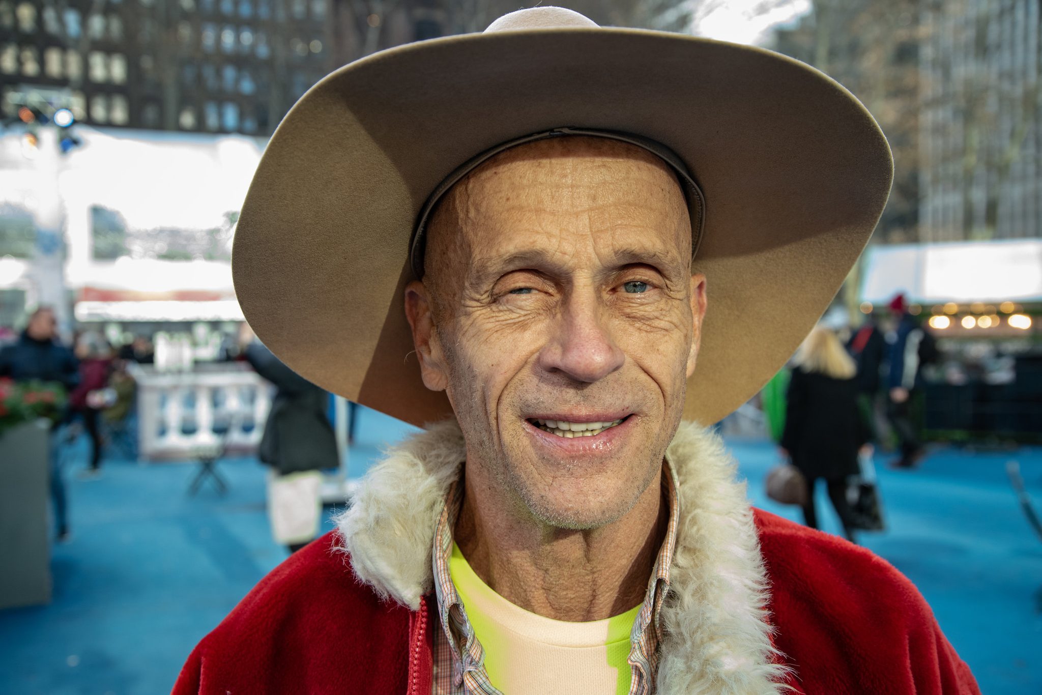 A man in a cowboy hat and a fur-collared coat grins for the camera beside the winter rink.
