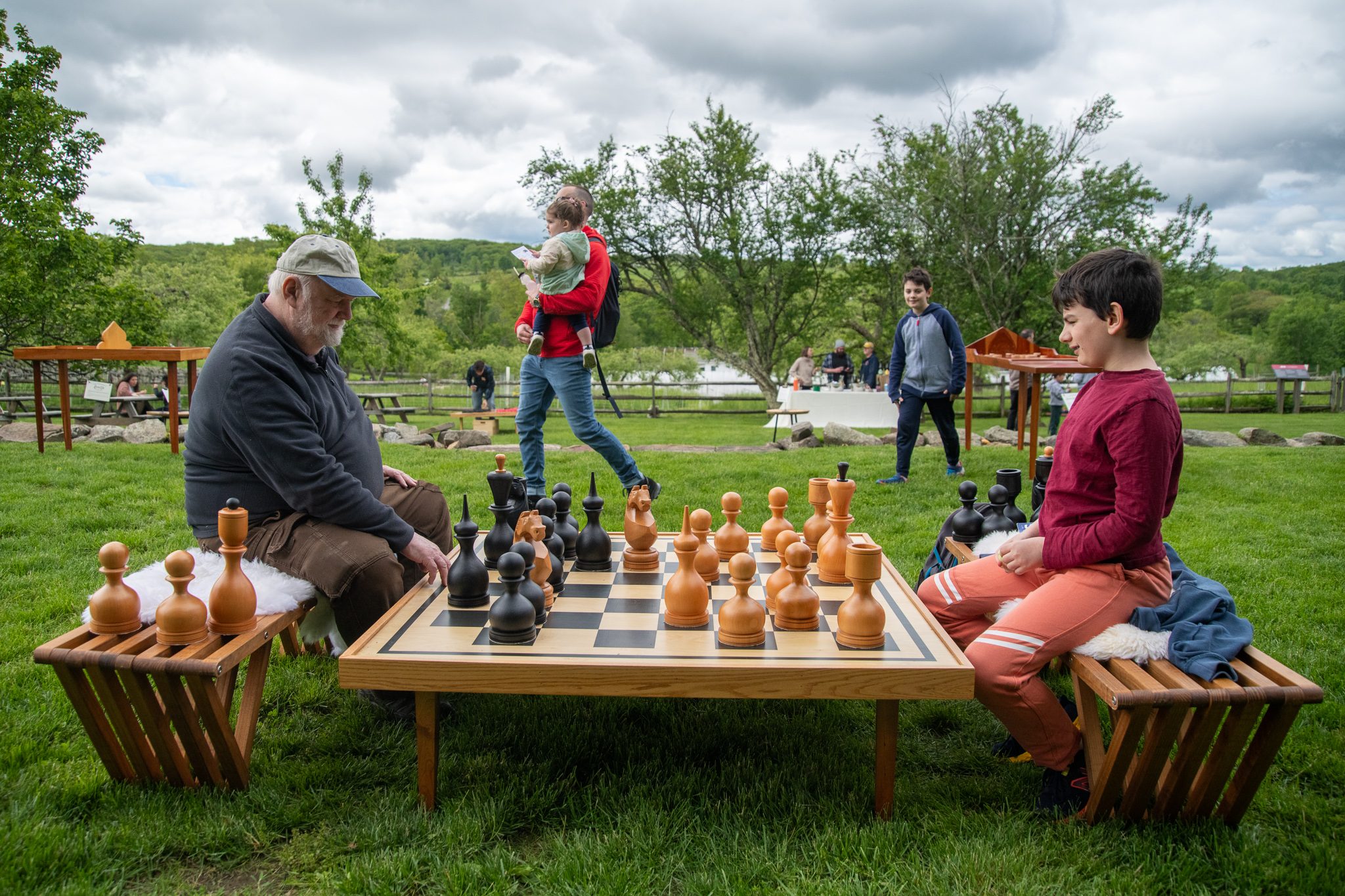An old hand and a young one face off over a giant chess board on the lawn.