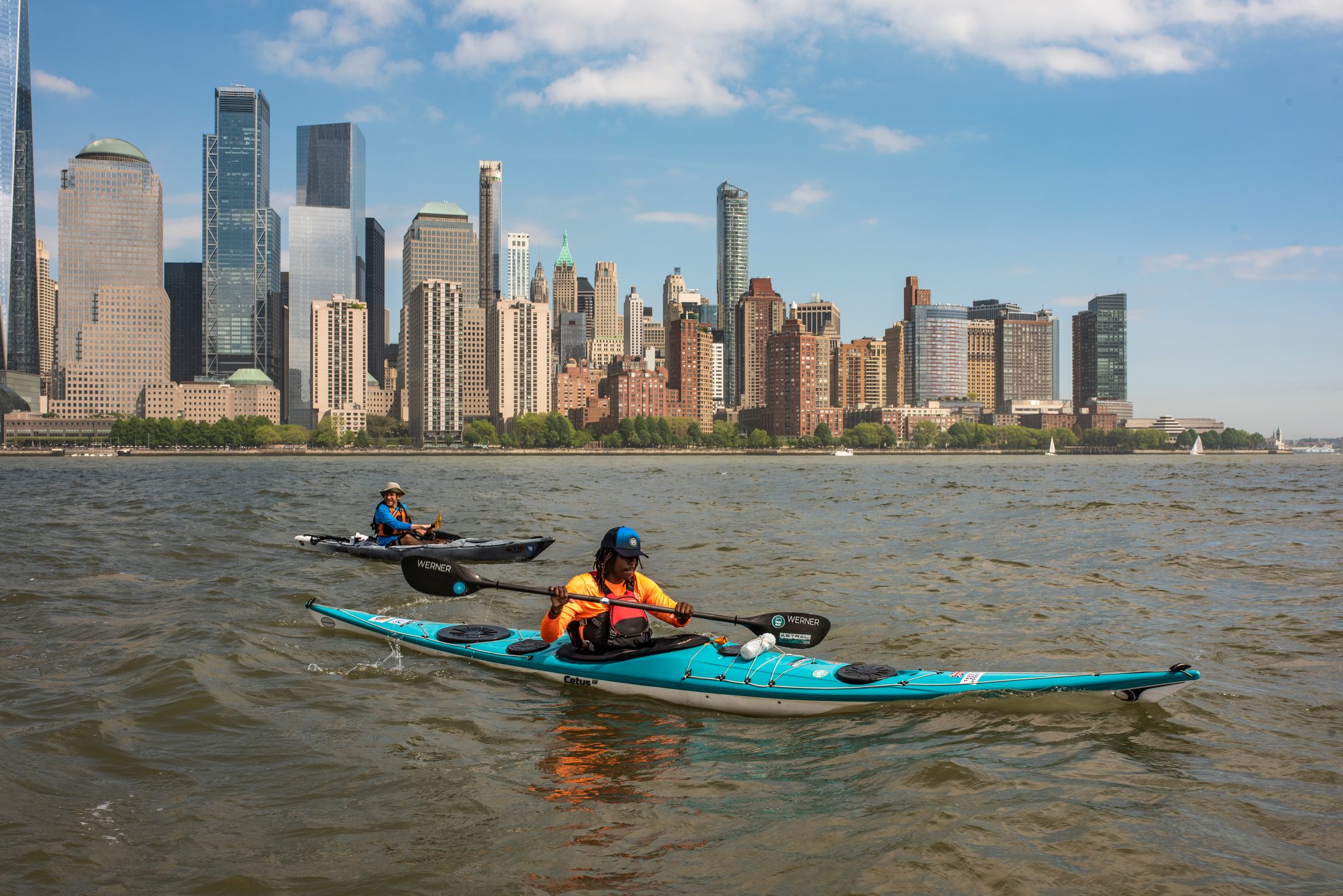 Orange jacket, teal kayak, the downtown skyline filling the whole horizon behind.