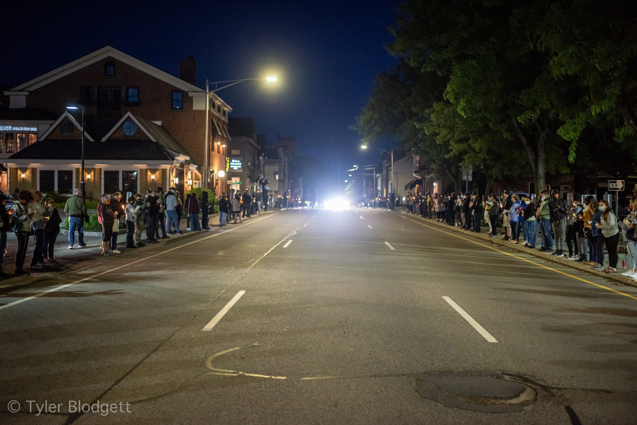 People line both sides of an empty street by candlelight, a single bright light waiting at the far end.