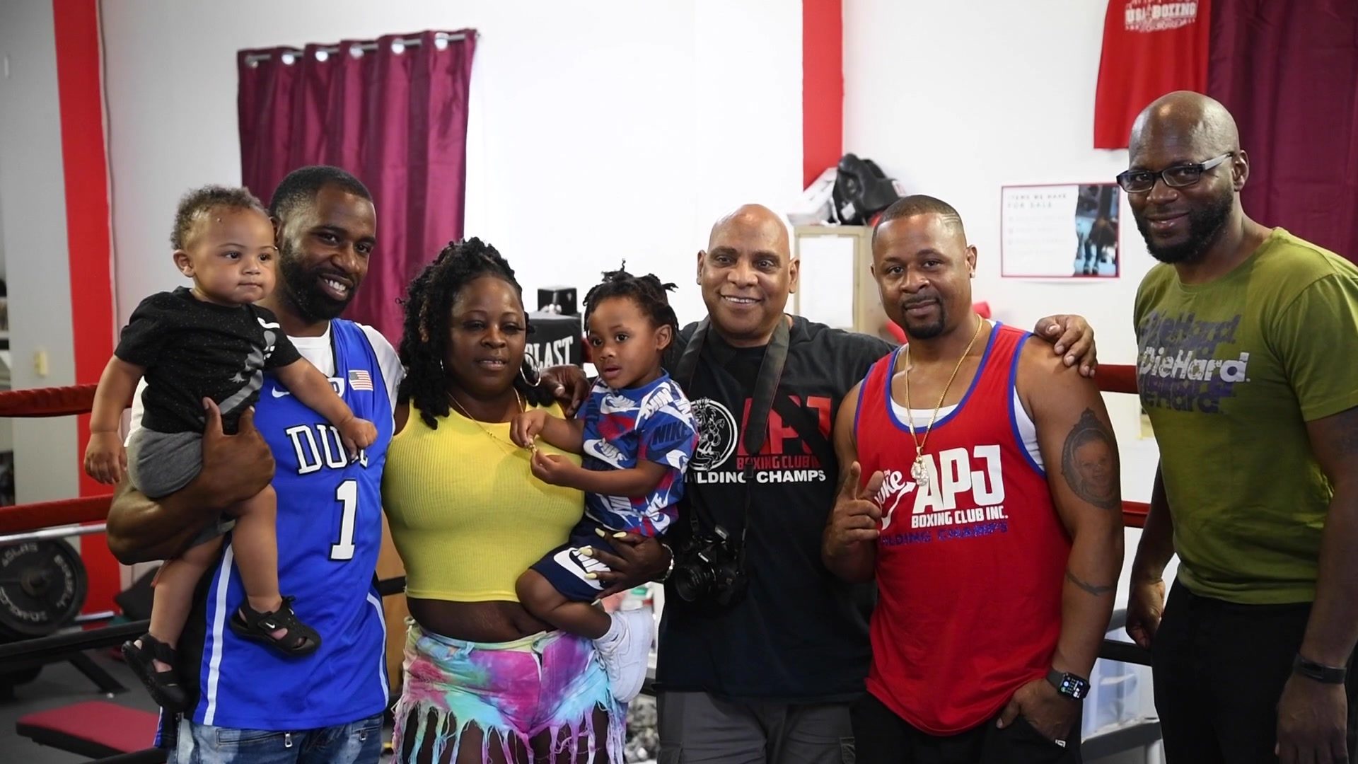 A group gathers for a photo by the ring, two toddlers held up in arms and a red club tank at center.