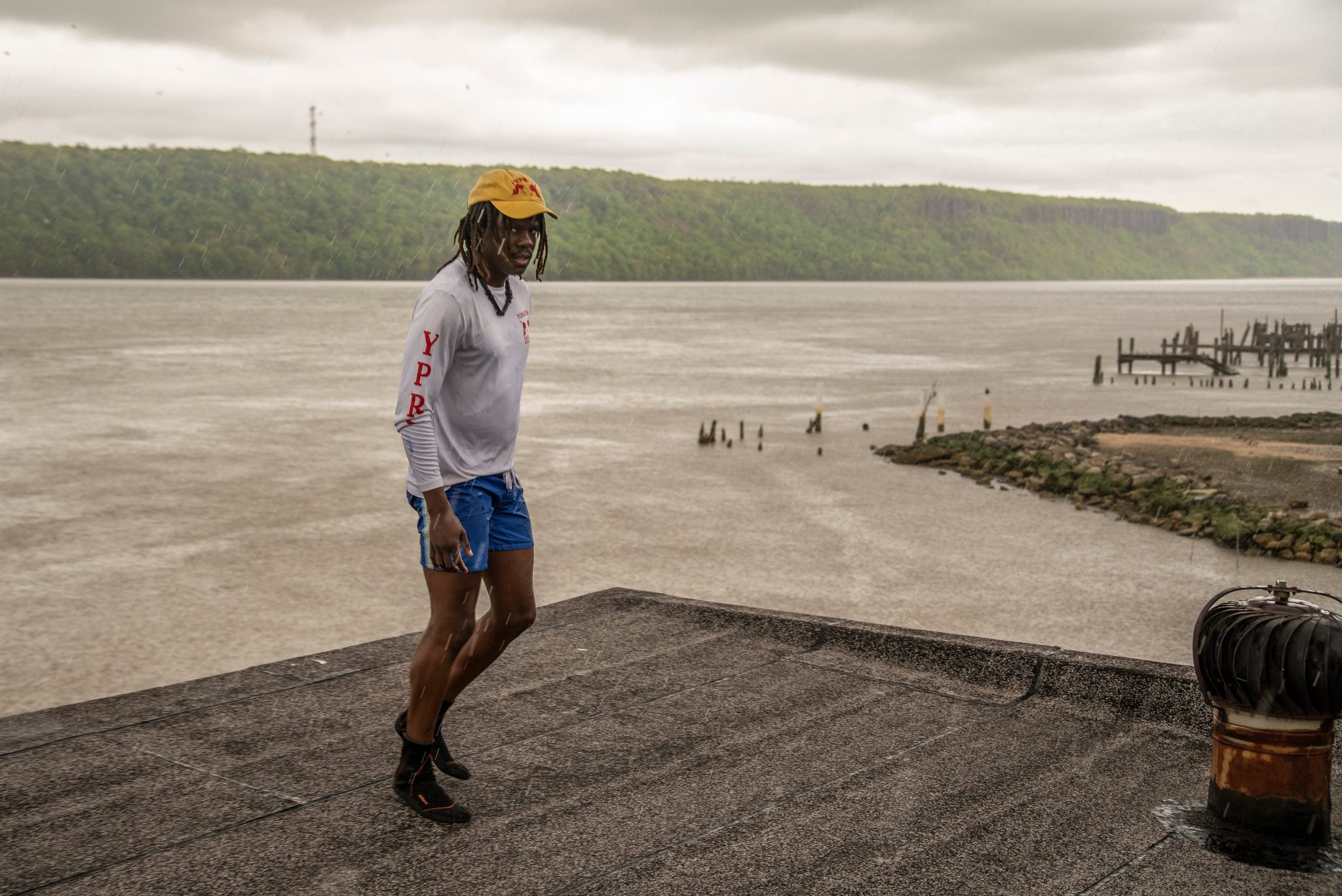 Chev walks the weathered dock in his club shirt, the river and the long ridge of cliffs behind him.