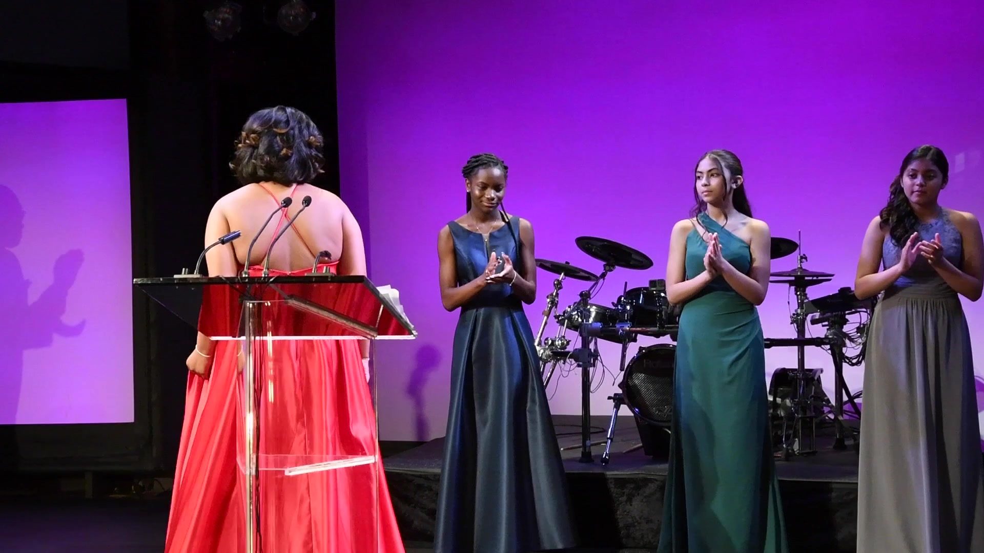 The young women stand in their gowns as a speaker addresses the room.