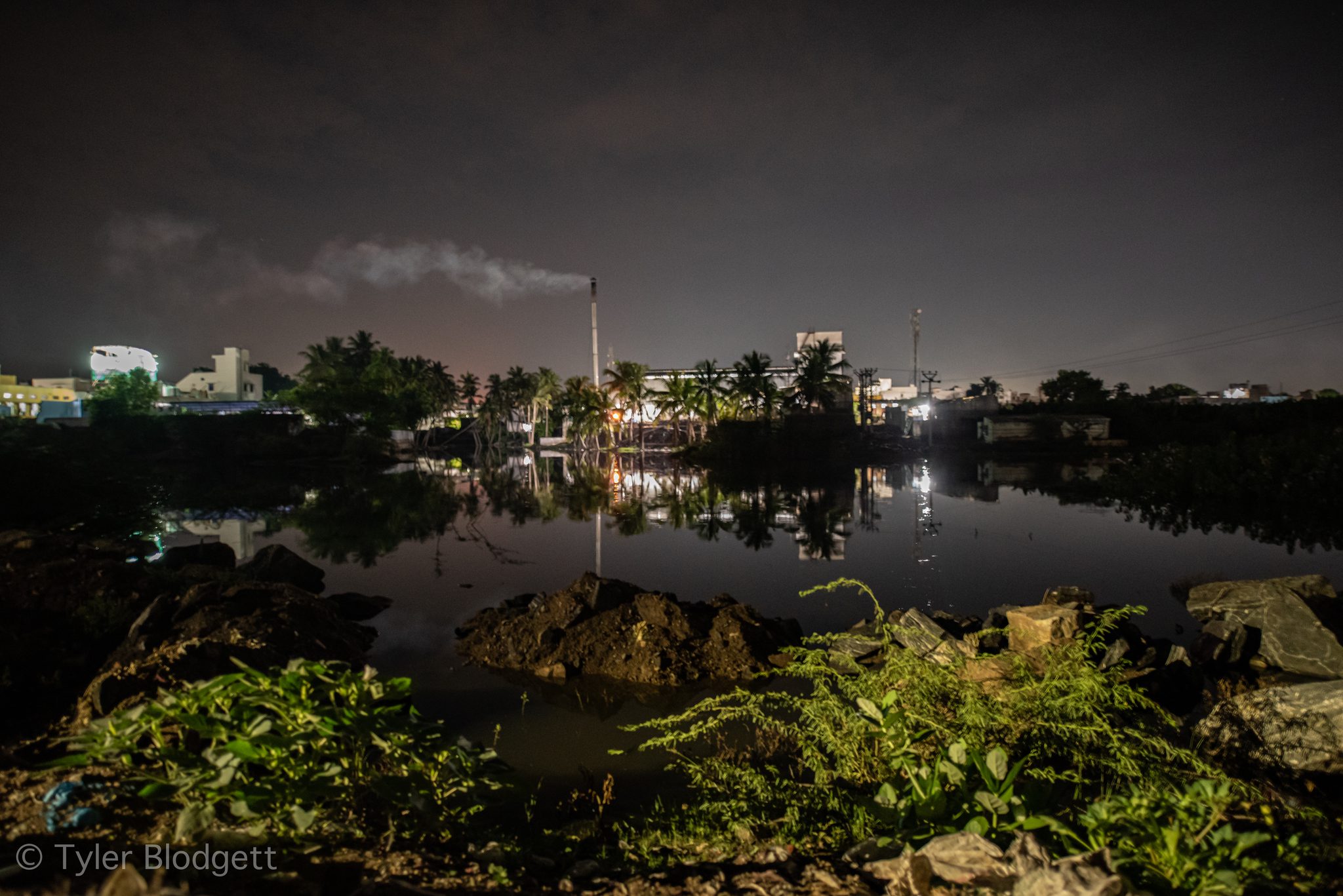 The lights of the town and a smoking chimney reflect in the still river at night.