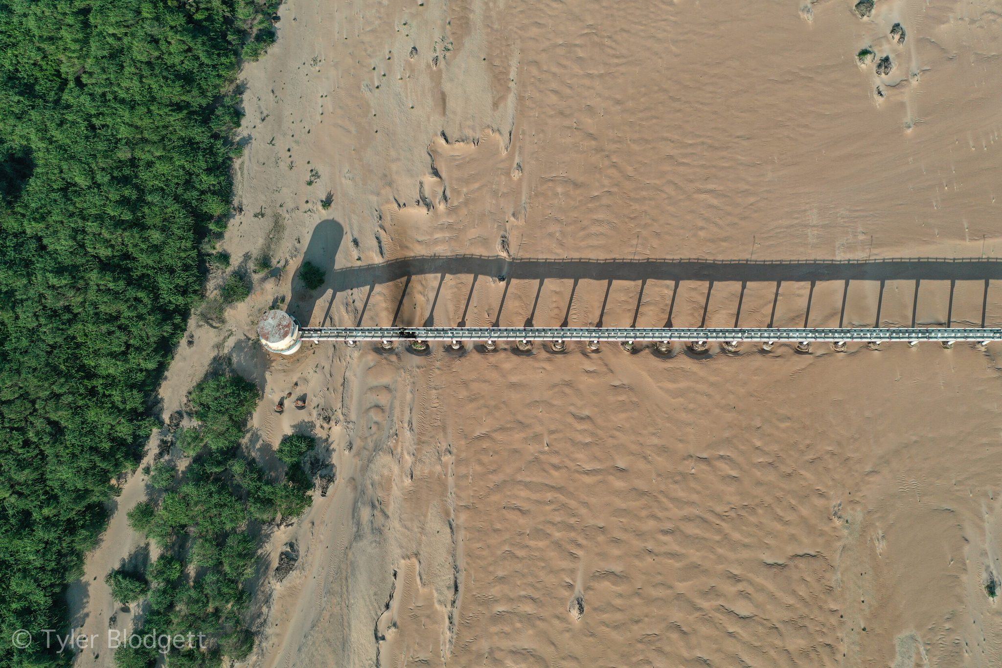 A narrow bridge lays its shadow across a riverbed of rippled sand.