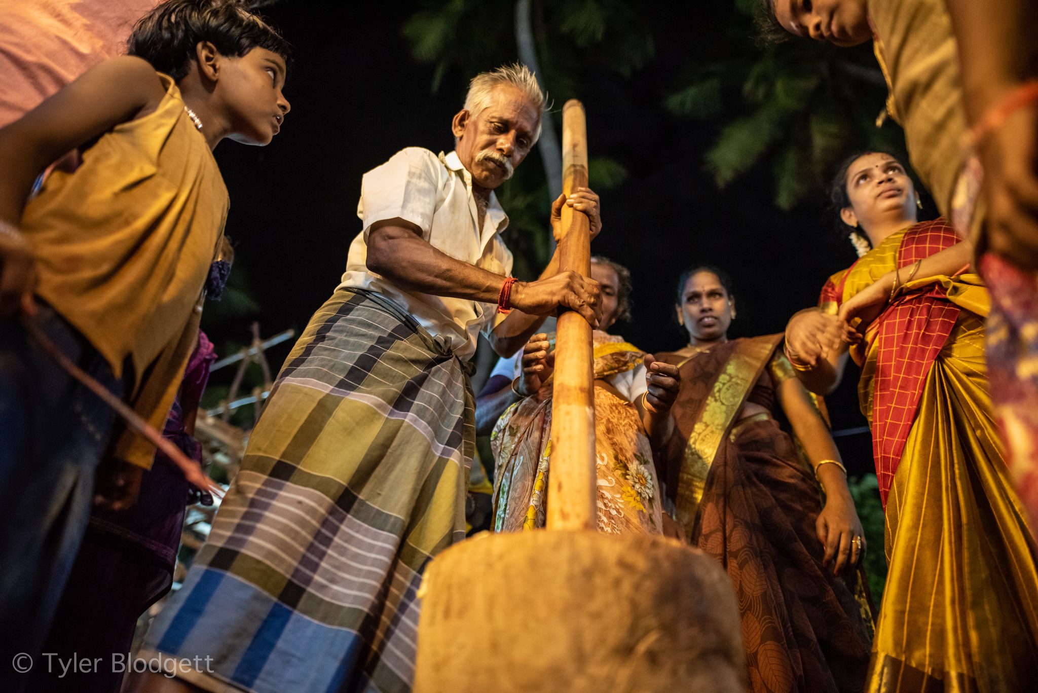 An elder grips the long wooden pestle as the family gathers close in the night.