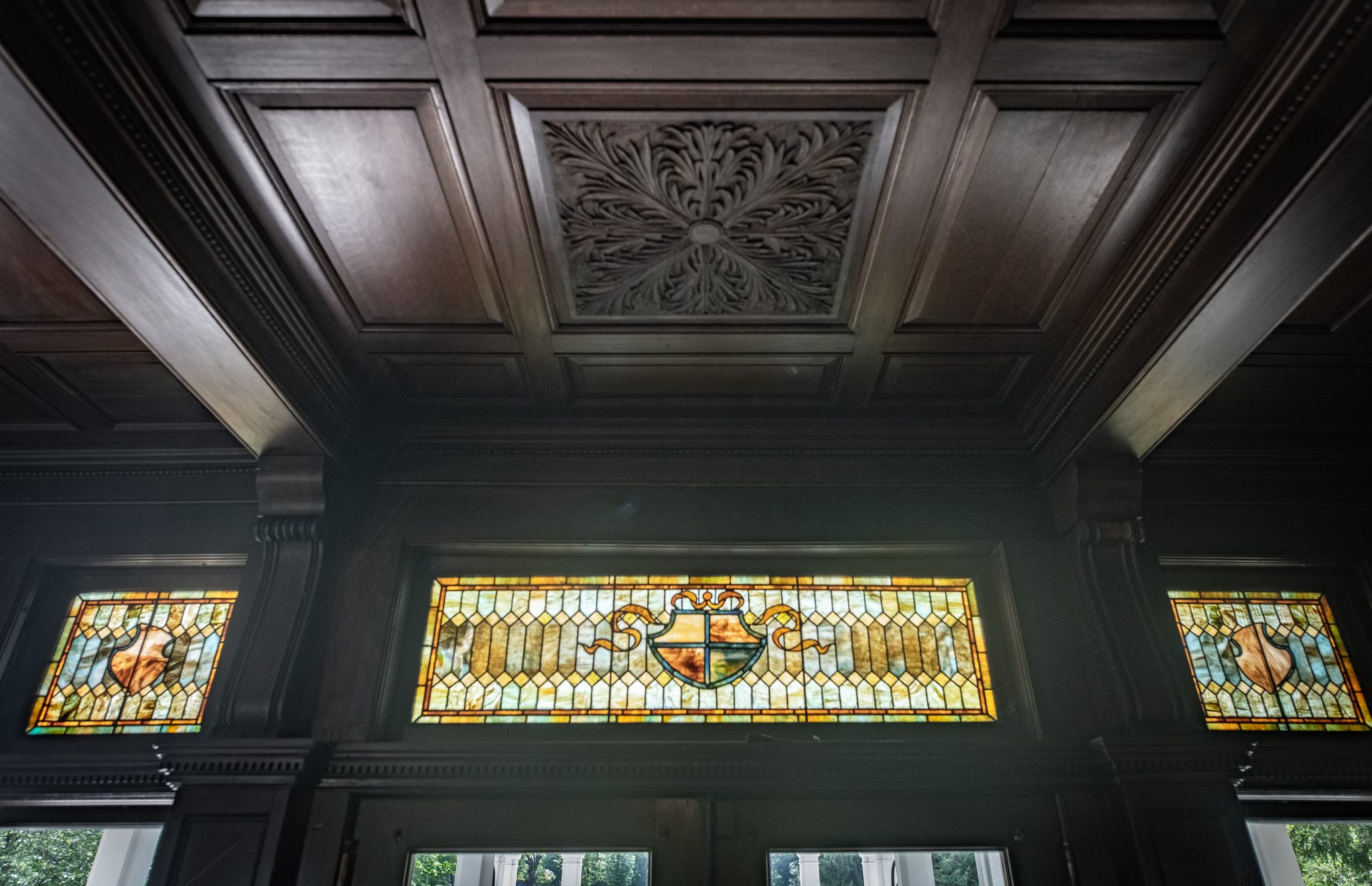 Looking up in the same paneled room, a carved coffered ceiling above a row of stained-glass transoms with painted shields.
